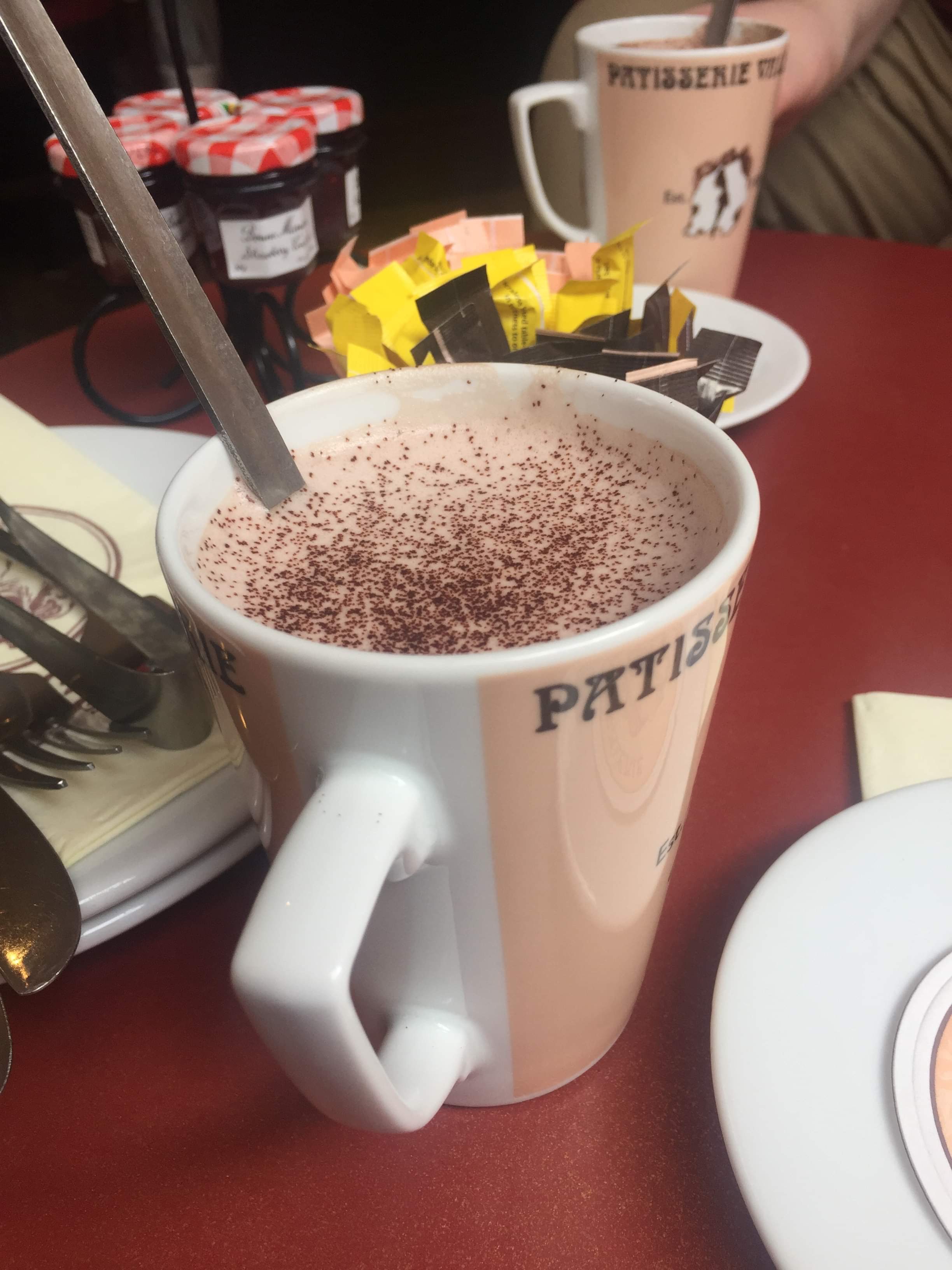 You've Got Chocolate All Around Your Mouth | Superdrug Skin Rescue Chocolate Orange Self-Heating Mask A tall brown and cream ceramic mug with a long silver metal spoon full of some dark brown hot chocolate and chocolate sprinkles on a dark wooden table, on a white background.
