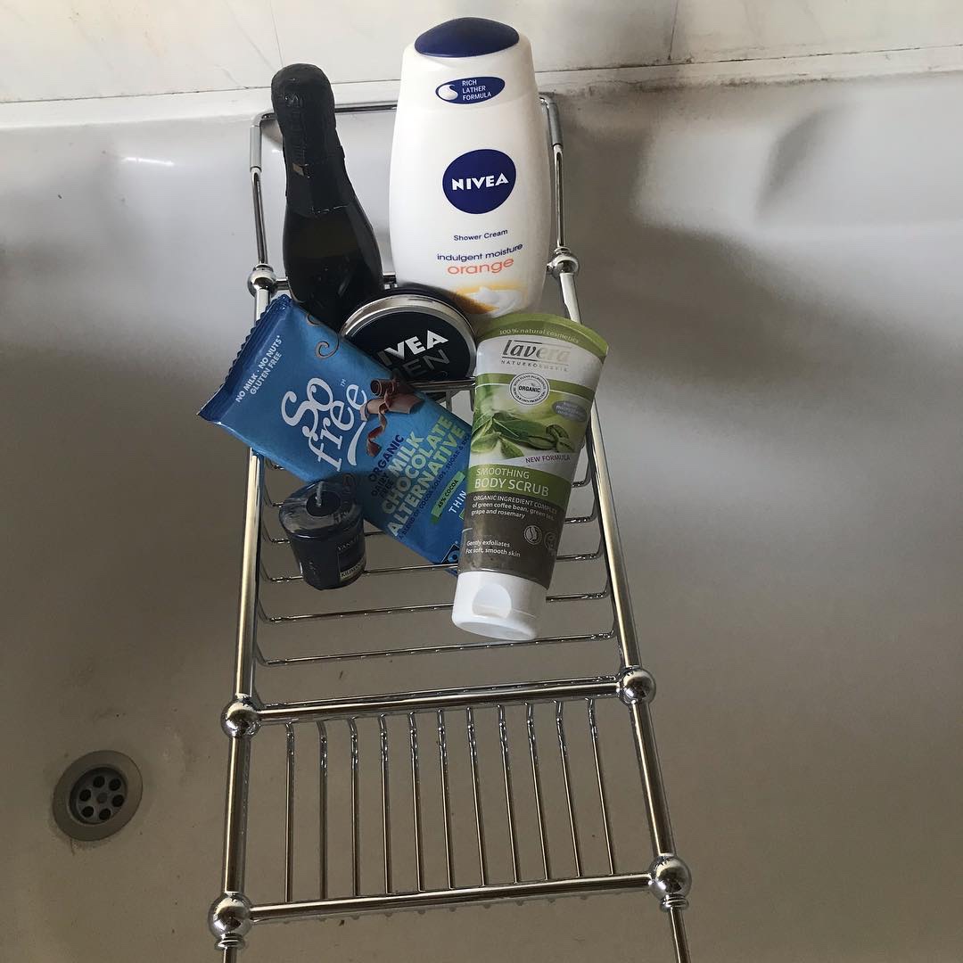 A tall rounded rectangular white plastic bottle that has Nivea Shower Cream Gel Indulgent Moisture Orange written in bold black writing on it, a tall cone shaped green plastic bottle that has Lavera Smoothing Body Scrub written in bold black writing on it, a circular black metal tin that has Nivea Cream written in bold white writing on it, a tall black glass bottle that has Raffaello Prosecco written in bold black writing on it, a rectangular blue bar of chocolate that has So Free Fair Trade Milk Chocolate Alternative written in bold black writing on it, and a small dark purple votive Candle on a silver metal rack inside of a white bathtub, on a white background.