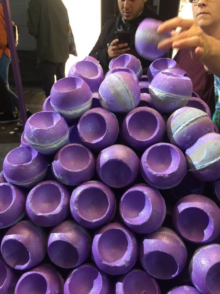A pyramid of some sparkly purple Bath Bombs that have holes at the top of them on a bright wooden stand, on a white background.