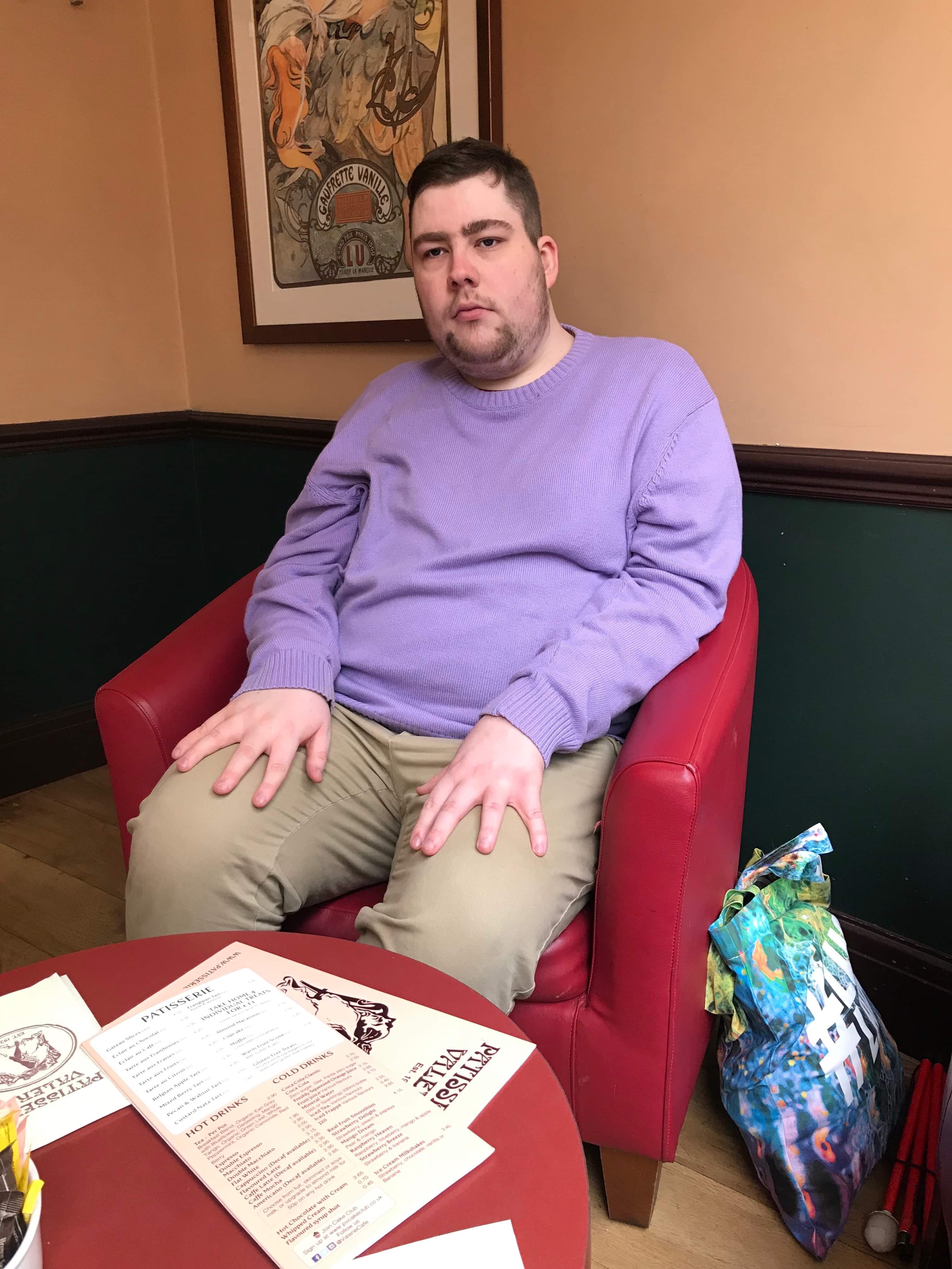 me sat on a red leather arm chair wearing a lilac jumper, a pair of navy chinos and a pair of black leather boots, on a white background.