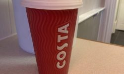 A tall cylindrical red ceramic cup with a white rubber lid that has costa engraved in bold white writing on it, on a white background.