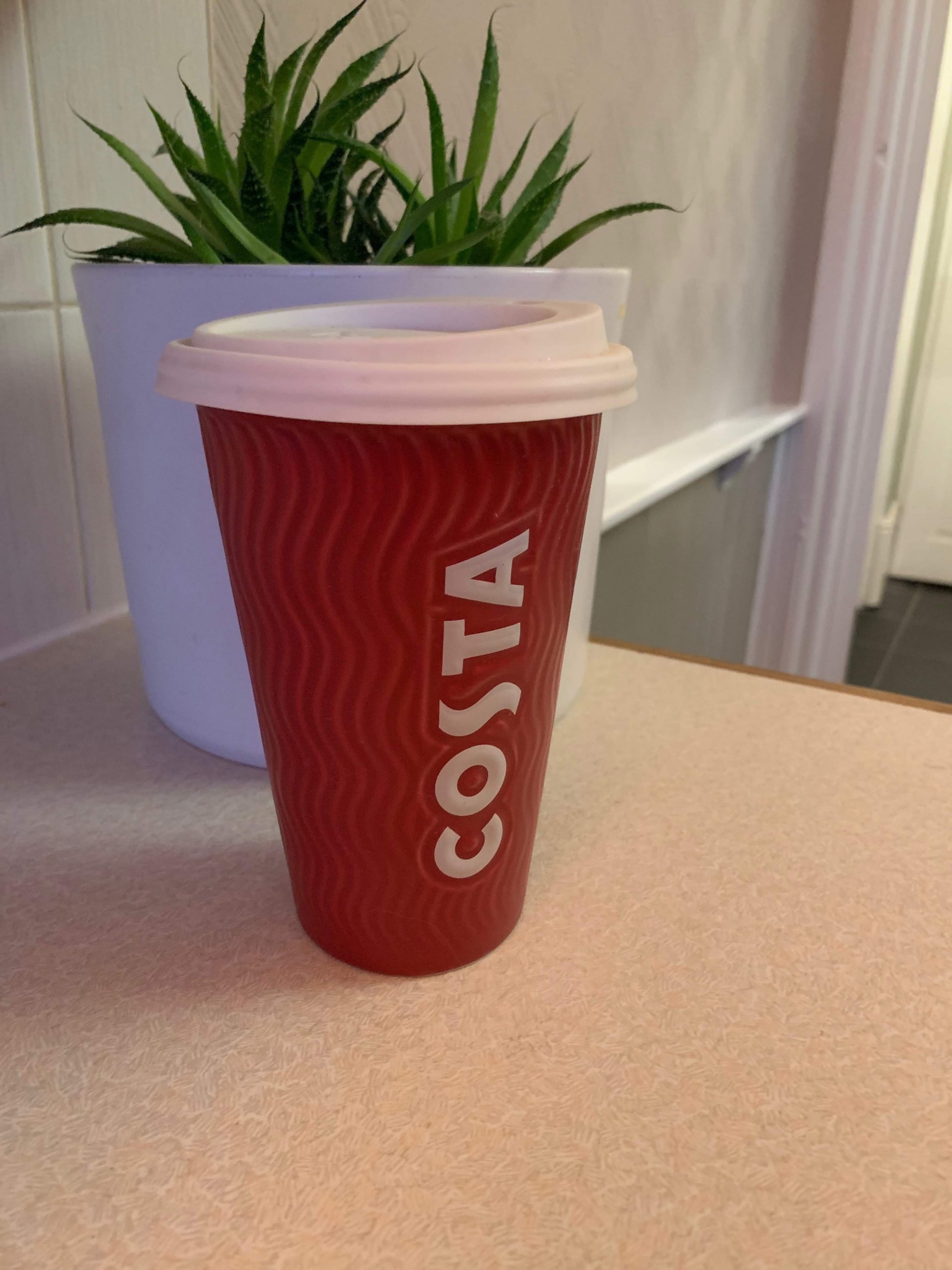 A tall cylindrical red ceramic cup with a white rubber lid that has costa engraved in bold white writing on it, on a white background.