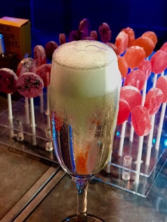 Sweet, Sugar, Candy Man! | Smith & Sinclair Leeds Pop Up Store Launch with The Alchemist Trinity Leeds A tall thin Champagne Flute full of some clear Prosecco, a cylindrical lolly pop and some white foam on a rectangular silver table covered in some blue cloth, on a dark background.