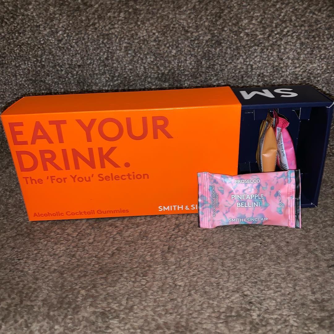 A large rectangular orange cardboard box full of some small rectangular plastic packets that has Eat Your Drink The For You Selection written in dark orange writing and Smith & Sinclair written in bold white writing on it, on a white background.