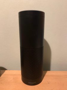 A tall cylindrical Black Plastic Speaker that has Amazon written in bold Silver writing on it, on a white background.
