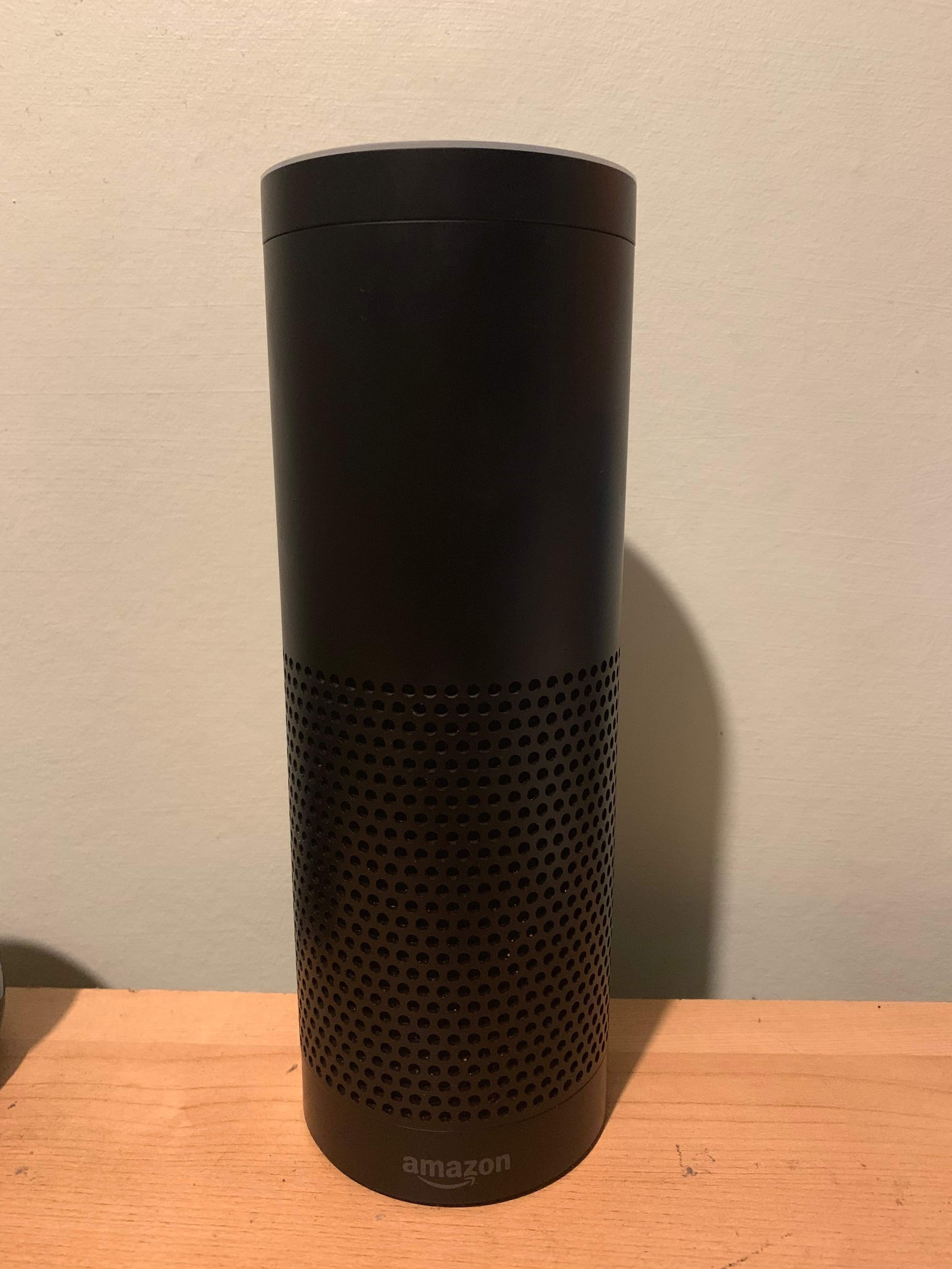 A tall cylindrical Black Plastic Speaker that has Amazon written in bold Silver writing on it, on a white background.