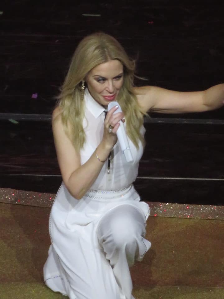 A short woman with blond hair wearing a white jumpsuit and holding a white microphone kneeling on one knee on a dark wooden stage, on a white background.