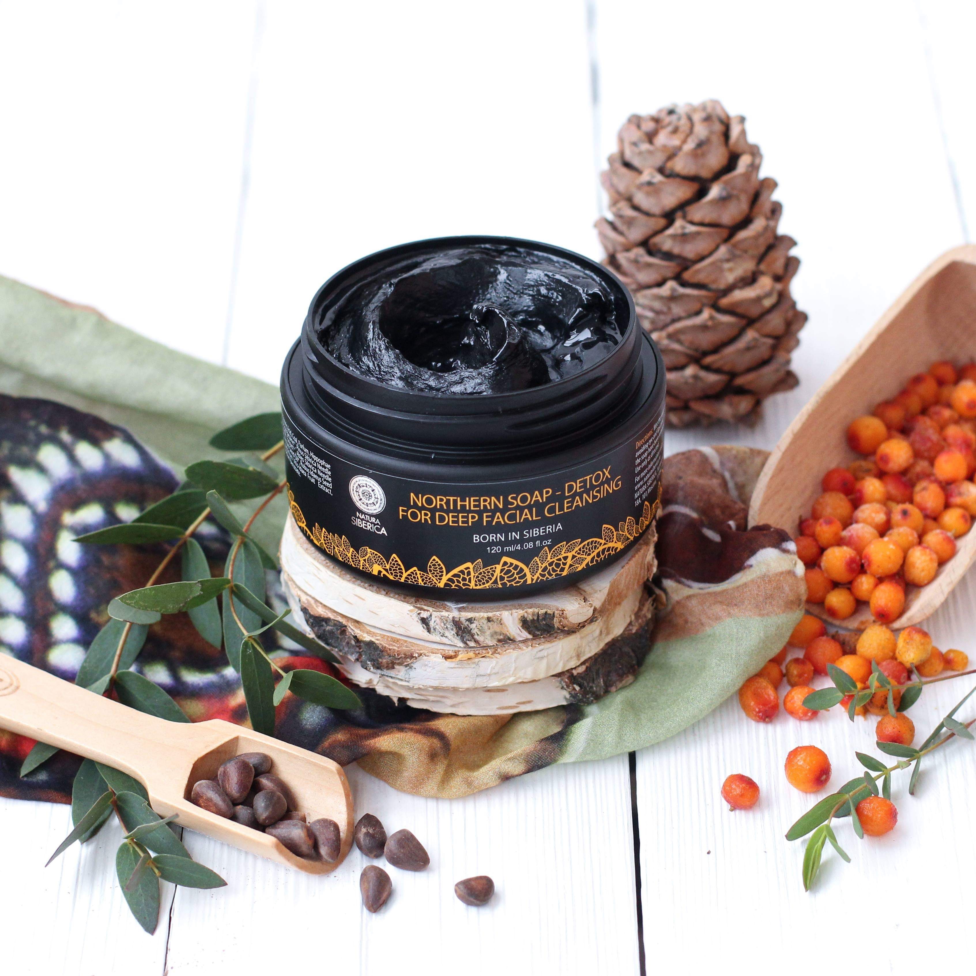 A short cylindrical black plastic tub with a black label that has northern soap for deep facial cleansing written in bright orange writing on it in front of some different coloured leaves and pinecones, on a white background.