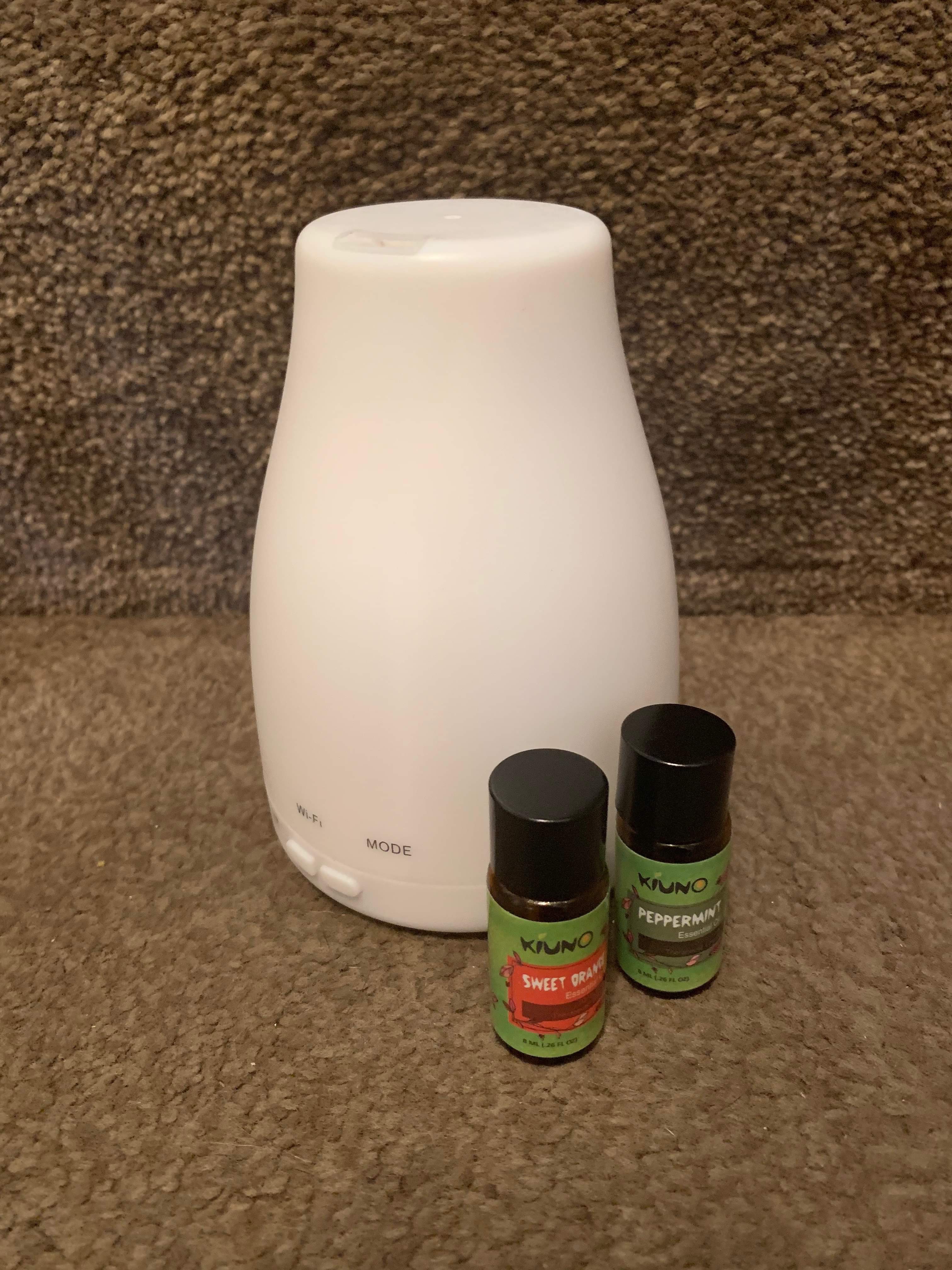 A tall conical shaped white Oil Diffuser with a hole at the top of it next to a brown glass bottle with a green label that has Kino Sweet Orange Oil written in bold white writing on it and a cylindrical brown glass bottle with a green label that has Kino Mint Oil written in bold white writing on it, on a white background.