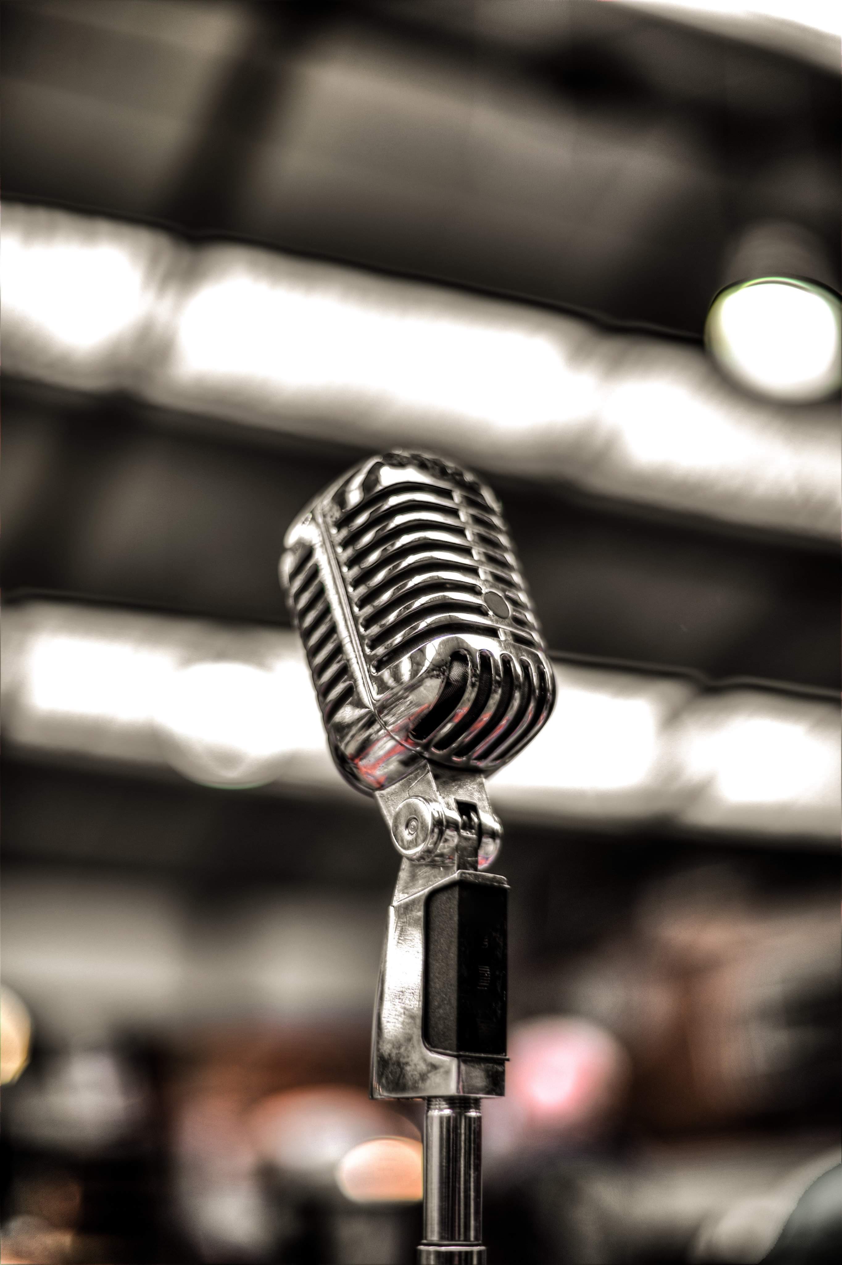 An old style Silver Microphone on a tall silver metal stand in front of a soft focus light wooden stage, on a white background.