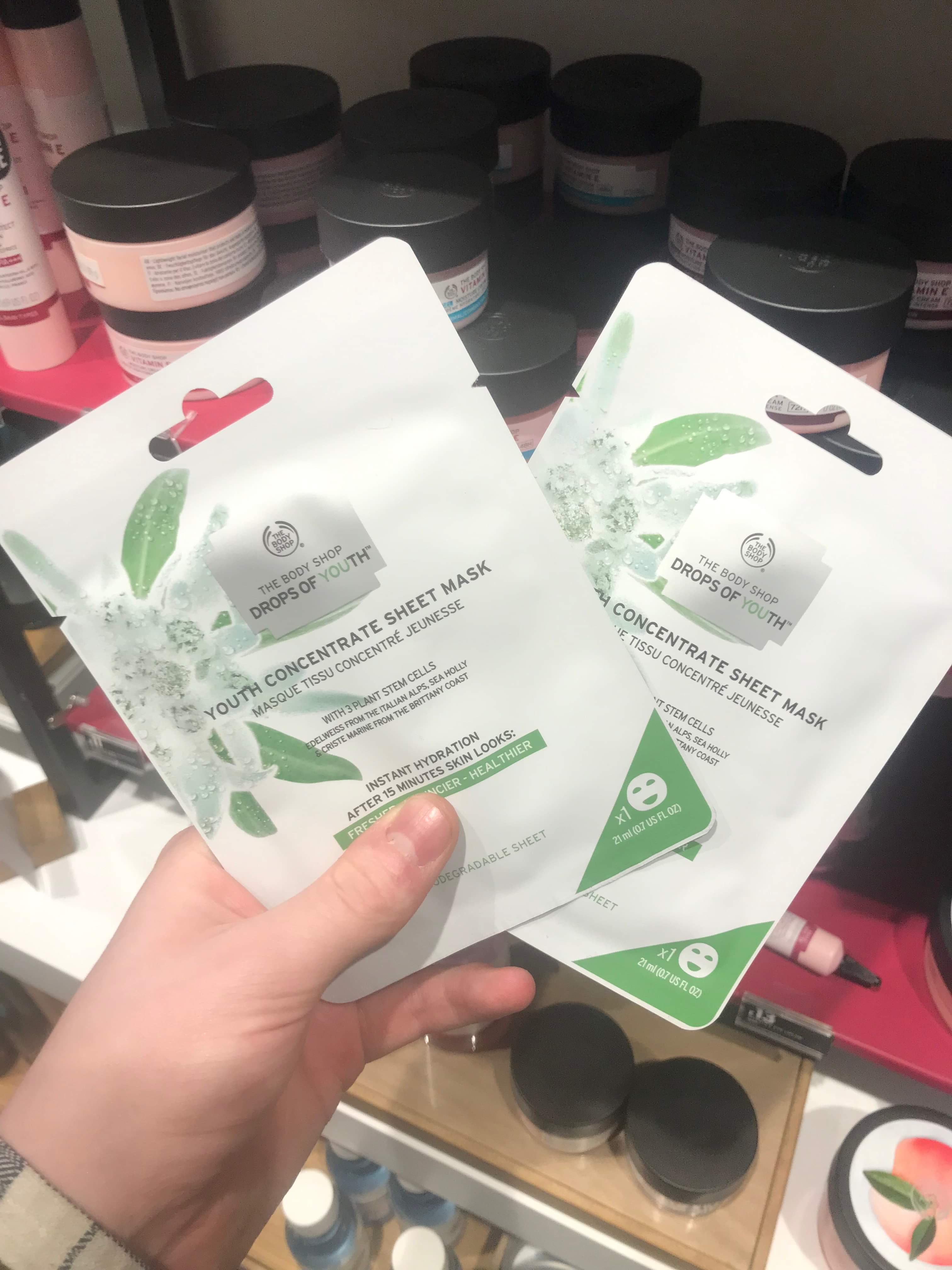 A hand holding two tall rectangular white plastic pouches that has The Body Shop Drops of Youth™ Youth Concentrate Sheet Mask written in bright green writing on it, on a white background.