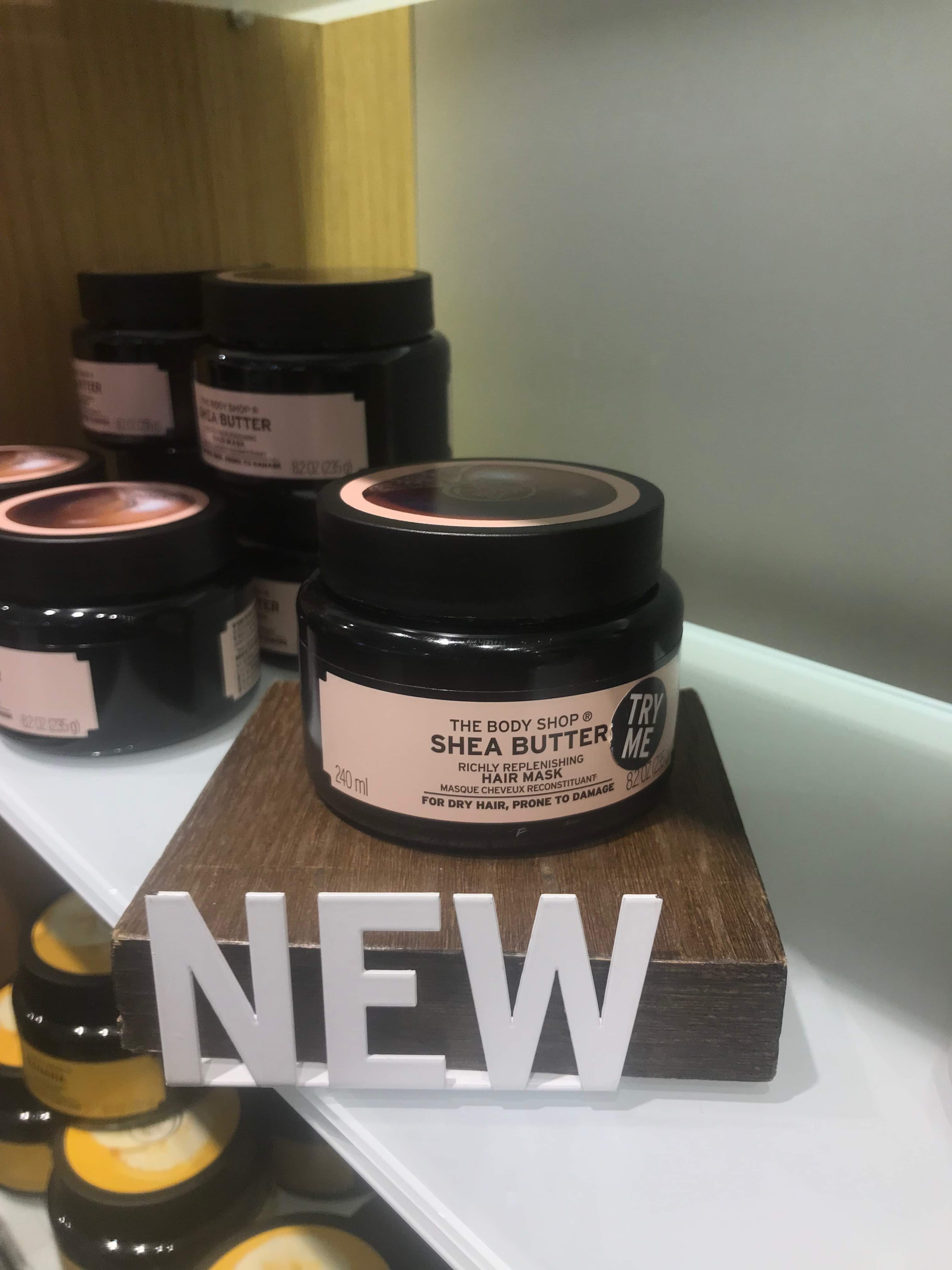 A short cylindrical Black Plastic Tub with a white label that has The Body Shop Shea Butter Richly Replenishing Hair Mask written in bold white writing on it, on a white background.