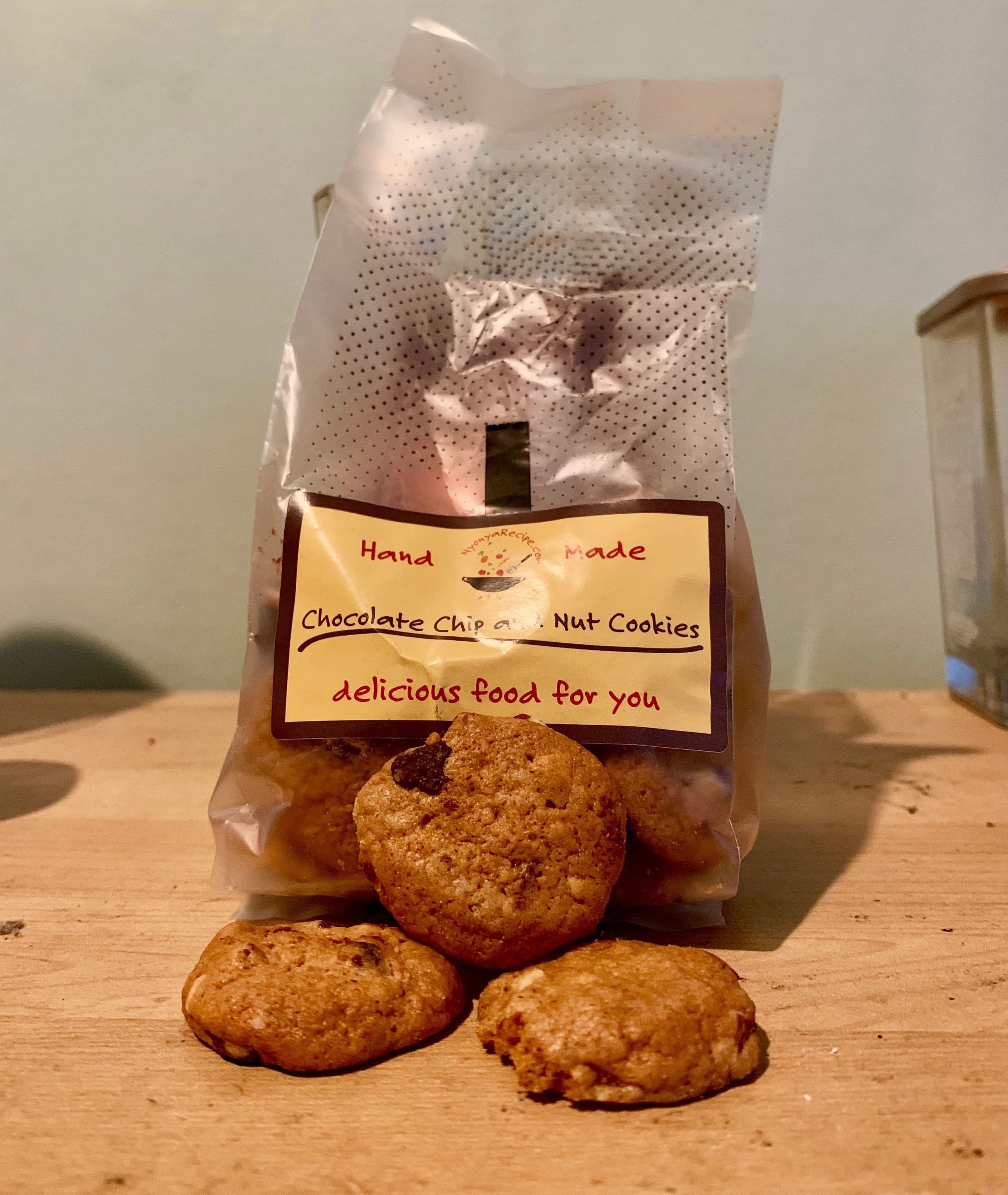 Eating My Way Through Malaysia | Nyonya Recipe Shop | Gifted Some small circular light golden brown cookies that have some Chocolate Chips embedded in them in front of a large rectangular clear Plastic Bag with a yellow label that has nyonya recipe shop chocolate chip & nut cookies written in bold red writing on it on a light wooden table, on a white background.