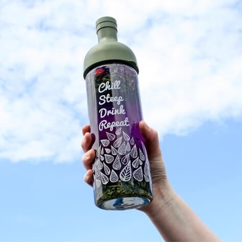 A tall cylindrical clear glass wine bottle with a green rubber neck and stopper that has chill, steep, drink , repeat written in bold white writing and some pictures of some White Leaves on it, on a white background.