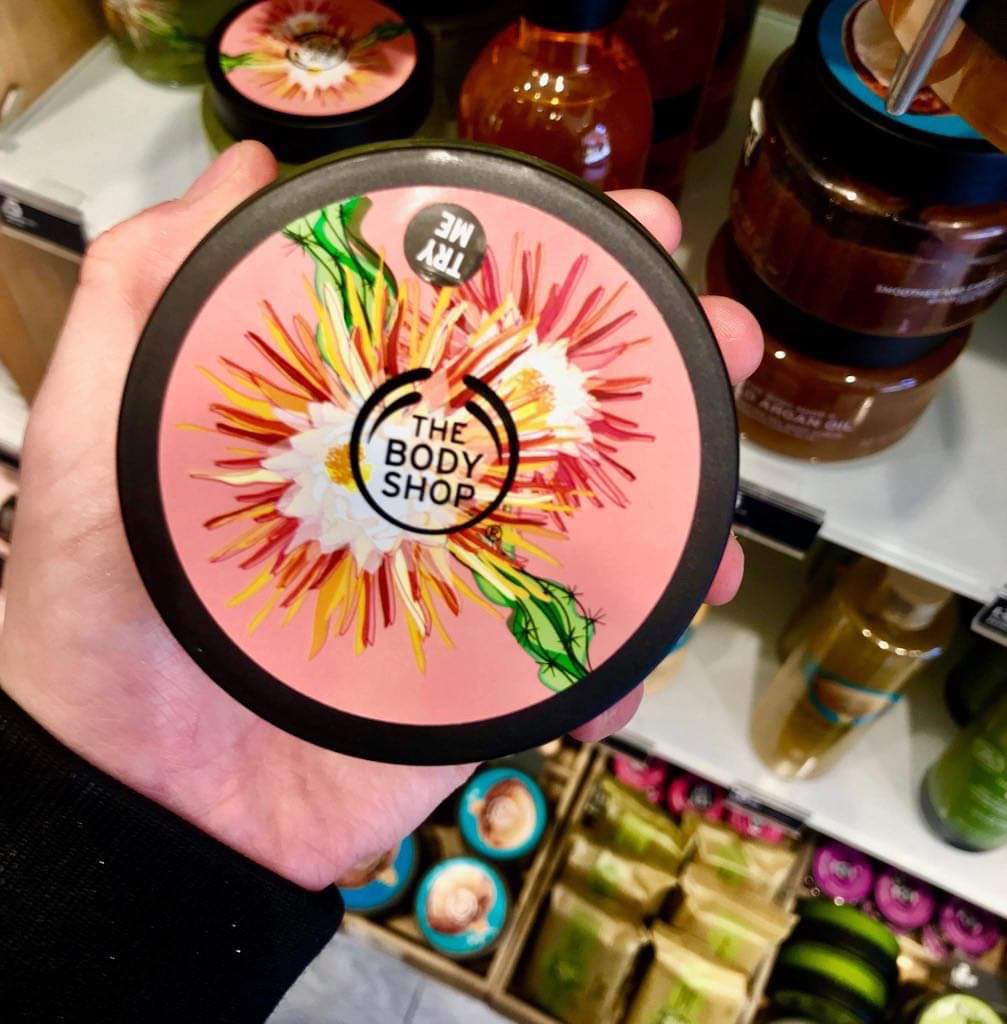 A short cylindrical black plastic tub with a bright pink lid that has The Body Shop written in bold black writing, Cactus Blossom Body Scrub written in dark pink writing, and some pictures of some red and yellow cactus blossoms on it, on a white background.