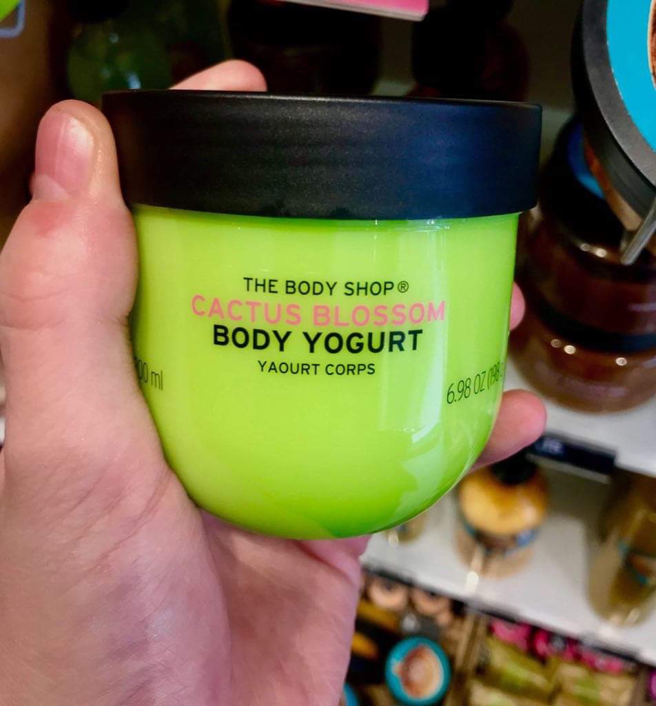 A short rounded rectangular green plastic tub with a black lid that has The Body Shop written in bold black writing, Cactus Blossom Body Yoghurt written in dark pink writing, and some pictures of some red and yellow cactus blossoms on it, on a white background.
