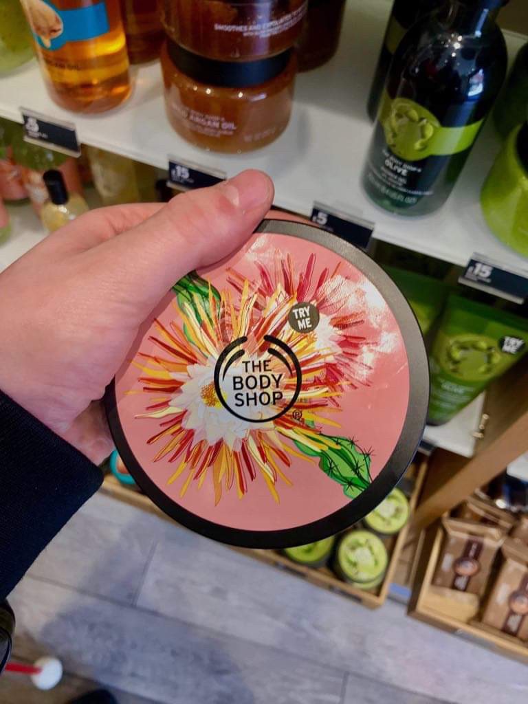 A short cylindrical black plastic tub with a bright pink lid that has The Body Shop written in bold black writing, Cactus Blossom Body Butter written in dark pink writing, and some pictures of some red and yellow cactus blossoms on it, on a white background.