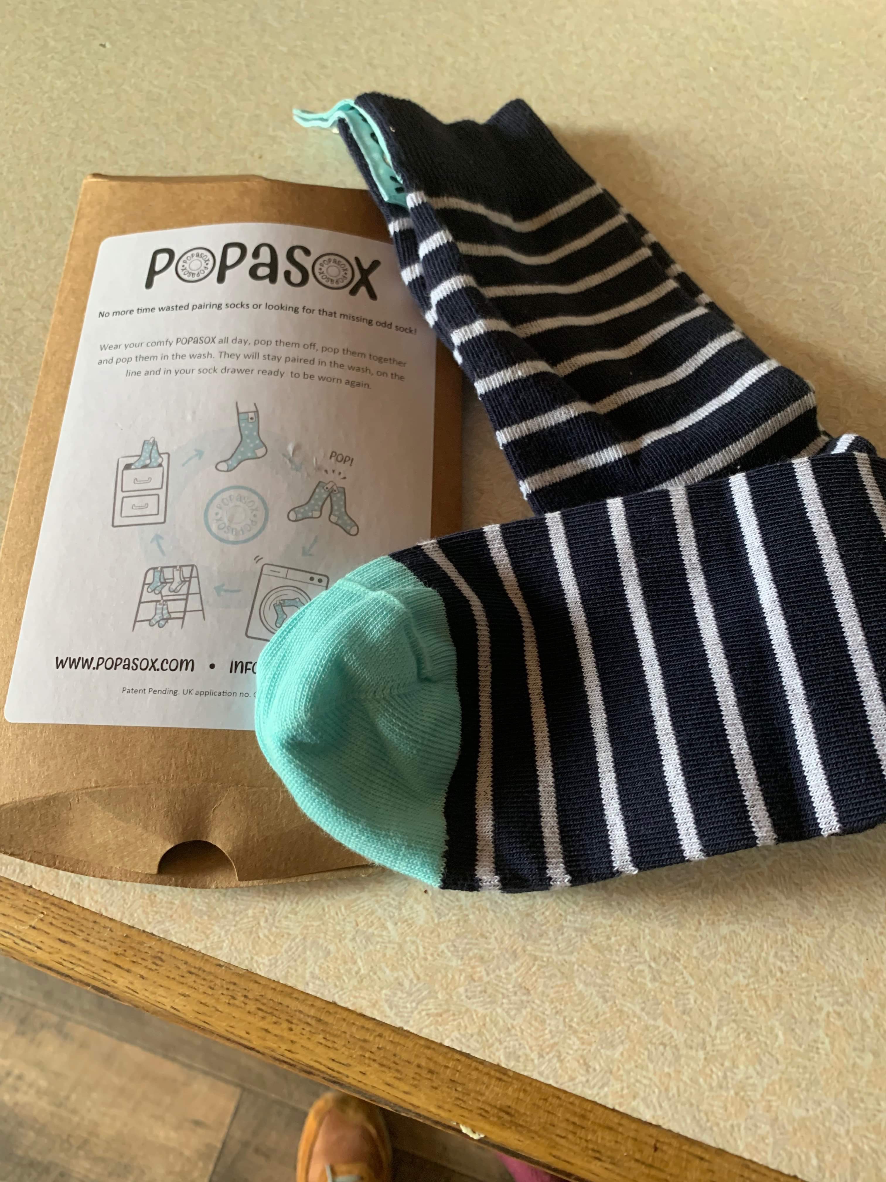 Pop Your Sox In The Wash | Popasox Men's Navy With White Stripe Socks | Gifted A blue and white striped sock that has a black popper and a bright green heel and toe next to a light brown cardboard sleeve with a white label that has Popasox Men's Navy with White Stripe Socks written in bold black writing on it, on a white background.