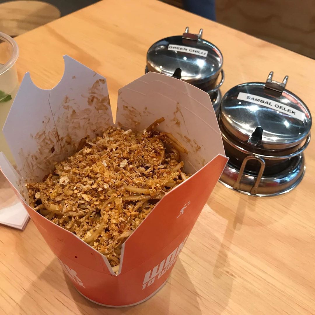 Walking Through Leeds With A Wok | Wok To Walk Leeds | Gifted A tall rectangular bright orange cardboard box full of some bright white egg noodles, some pieces of dark brown Beef, some crispy vegetables and some dark brown sauce on a light wooden table.