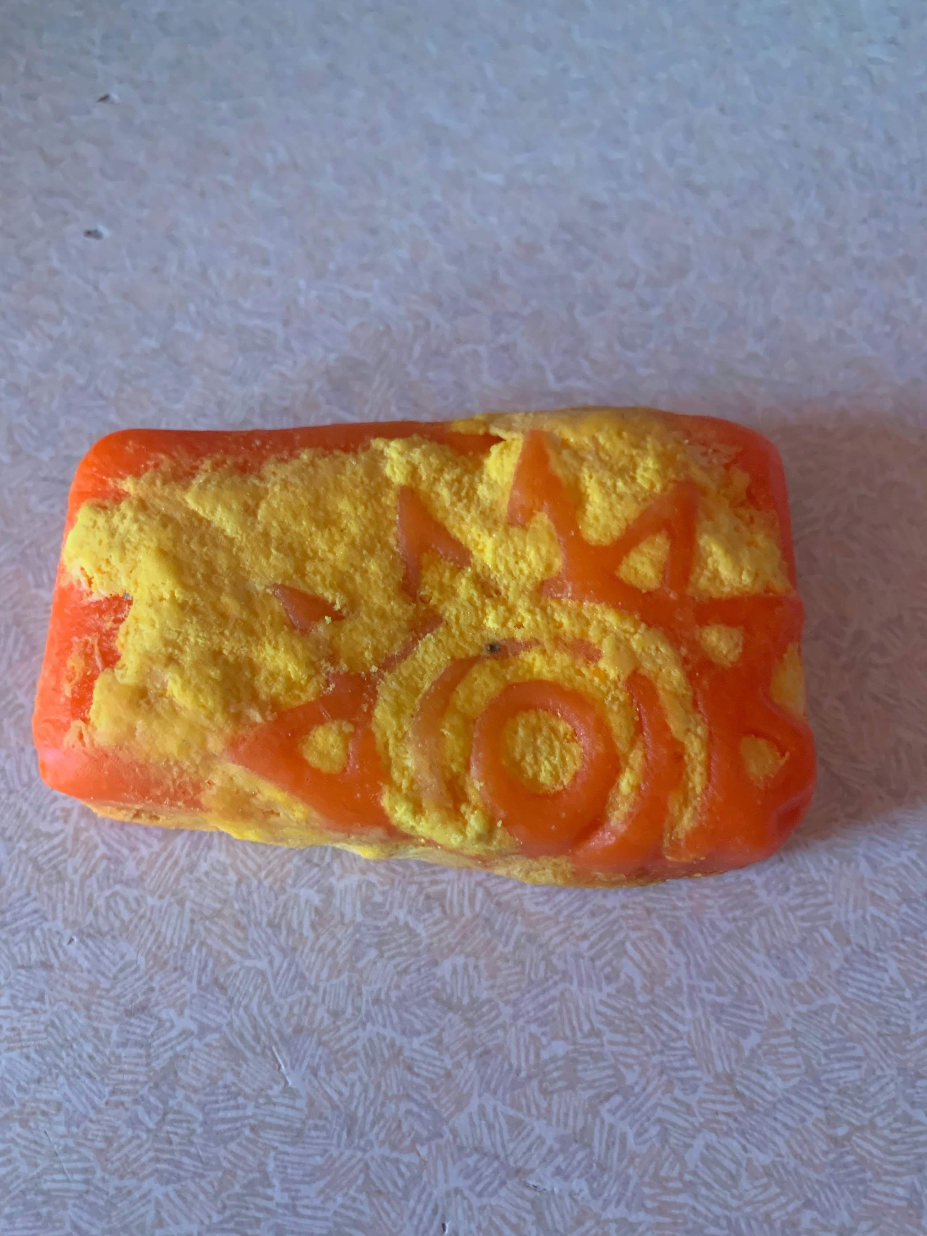 A large rectangular block of bright orange soap with a picture of a bright orange sun engraved on it that has a bright yellow outline, on a white background.