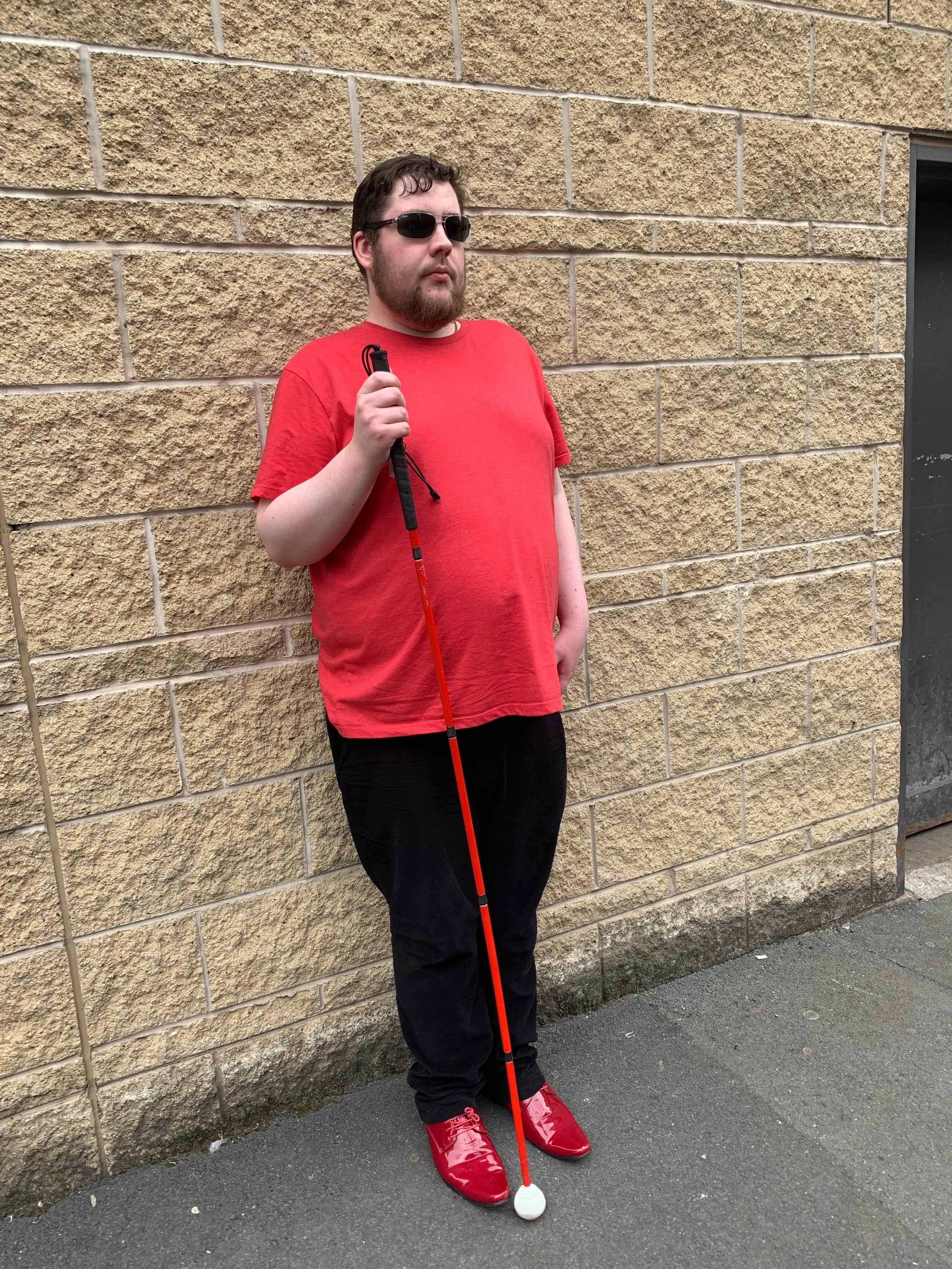 Outfit Of The Day | Cake'ole Bradford Me stood against a dark yellow brick wall wearing a pair of silver and black aviator sunglasses with some green lenses, a bright red crew neck t-shirt, a pair of black corduroy trousers, and a pair of shiny bright red shoes holding a long Red Cane, on a white background.