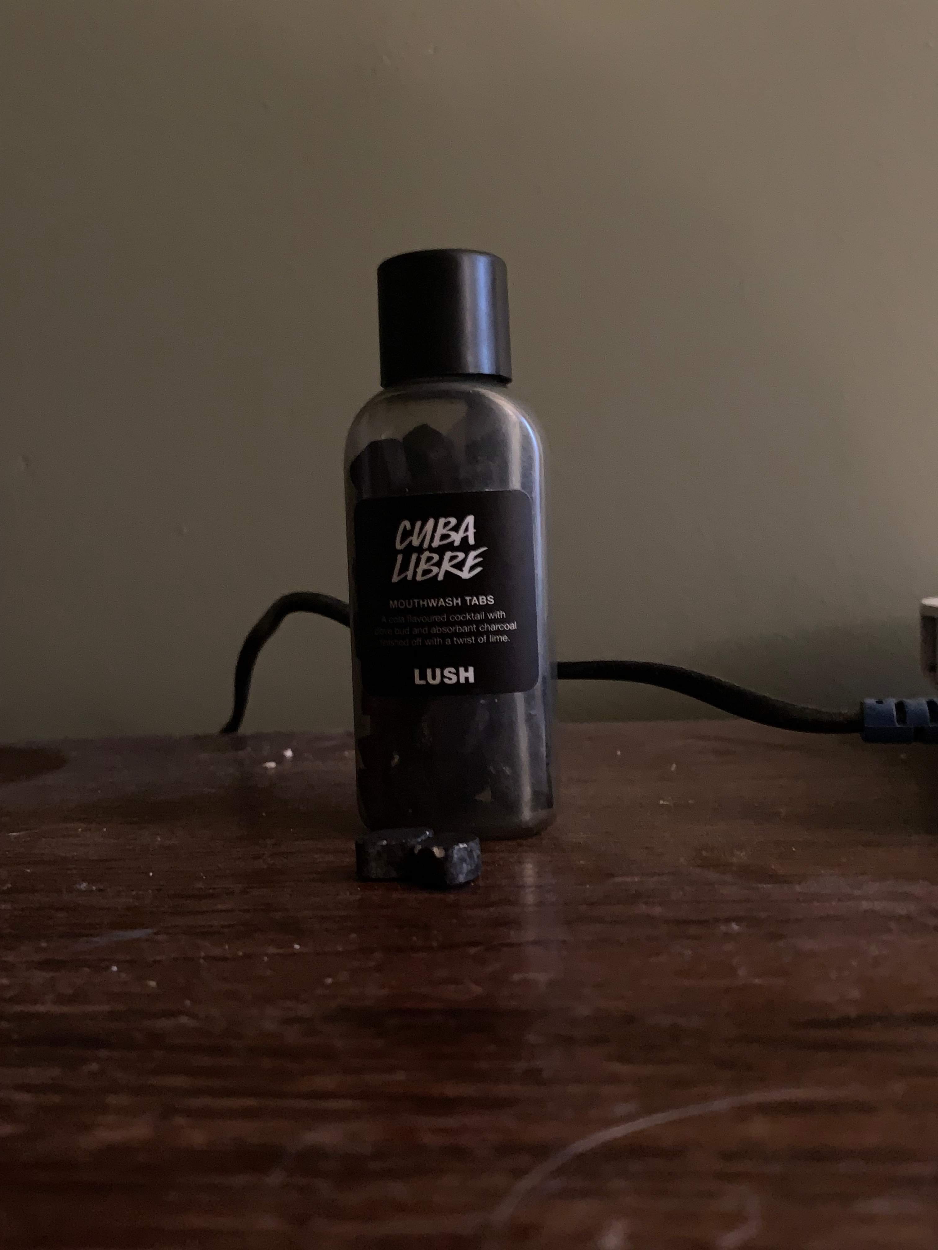 A short and flat rounded rectangular clear plastic bottle with a black lid and a black label that has Lush Cuba Libre Mouthwash Tabs written in bold white writing on it next to some small black diamond shaped pieces of powder, on a white background.