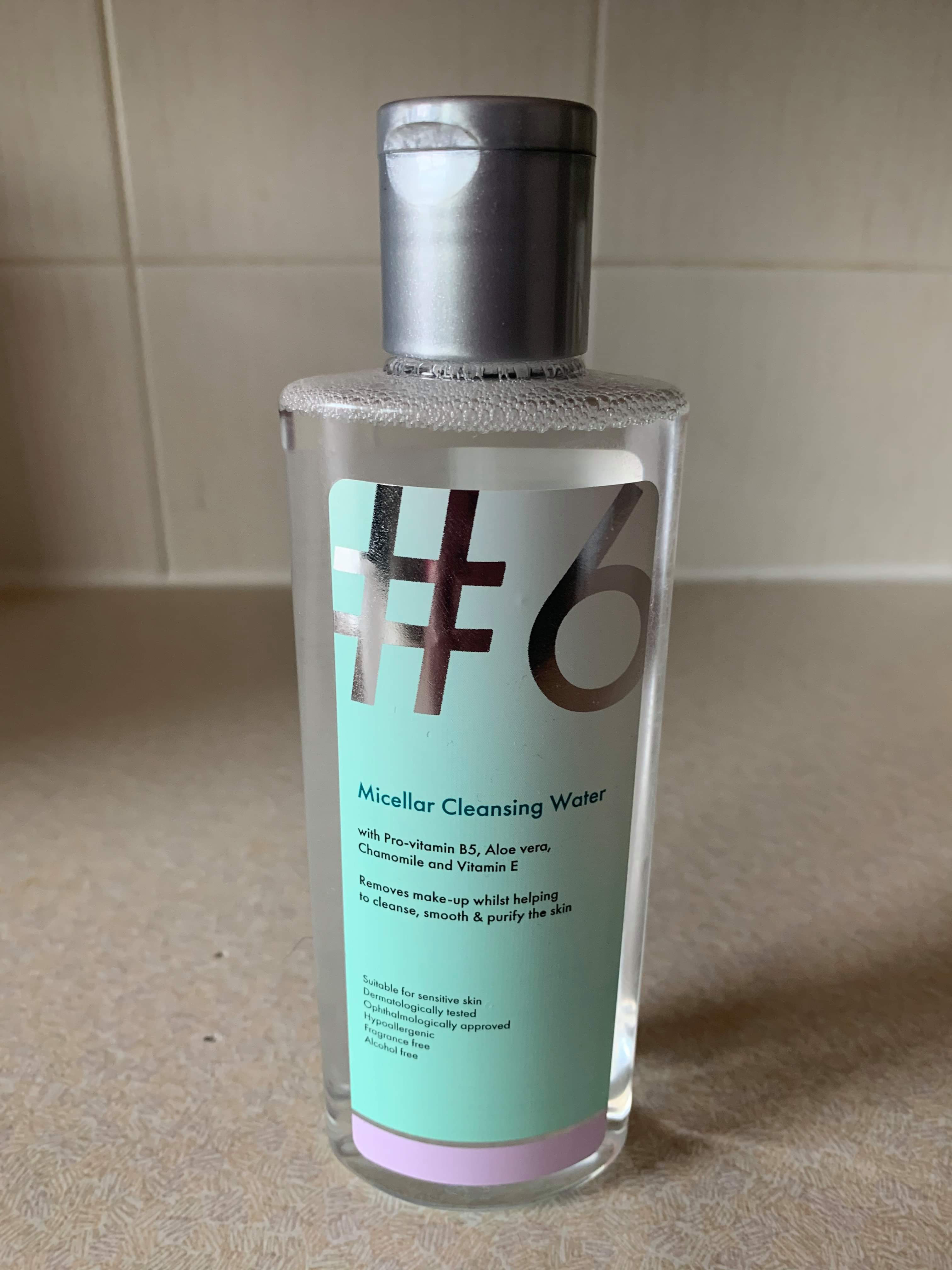 A tall rounded rectangular clear plastic bottle full of some clear liquid with a bright green lid and a bright green label that has Poundland #6 Micellar Cleansing Water written in bold black writing on it, on a white background.