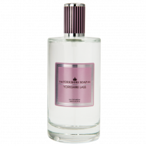 A tall cylindrical clear glass bottle full of some clear liquid with a silver and pink lid and a black label that has The Yorkshire Soap Company Yorkshire Lass Eau De Parfum written in bold pink writing on it, on a white background.