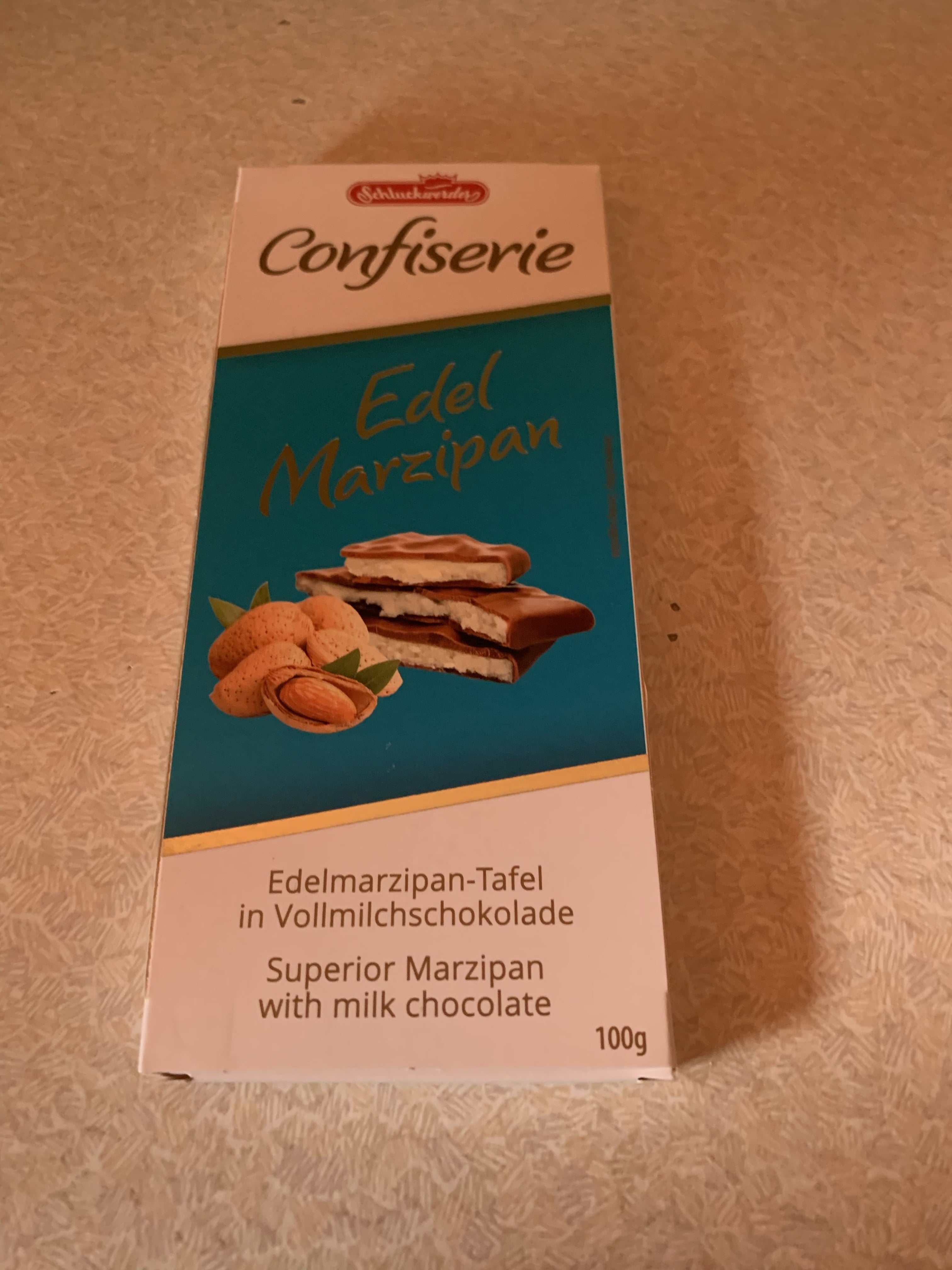 Marvellous Marzipan Magic | Lush Snowcake Soap and Snow Angel Bath Melt A tall rectangular blue and white Cardboard Box that has Schluckwerder Edel Marzipan with Milk Chocolate written in bold gold writing on it, on a white background.