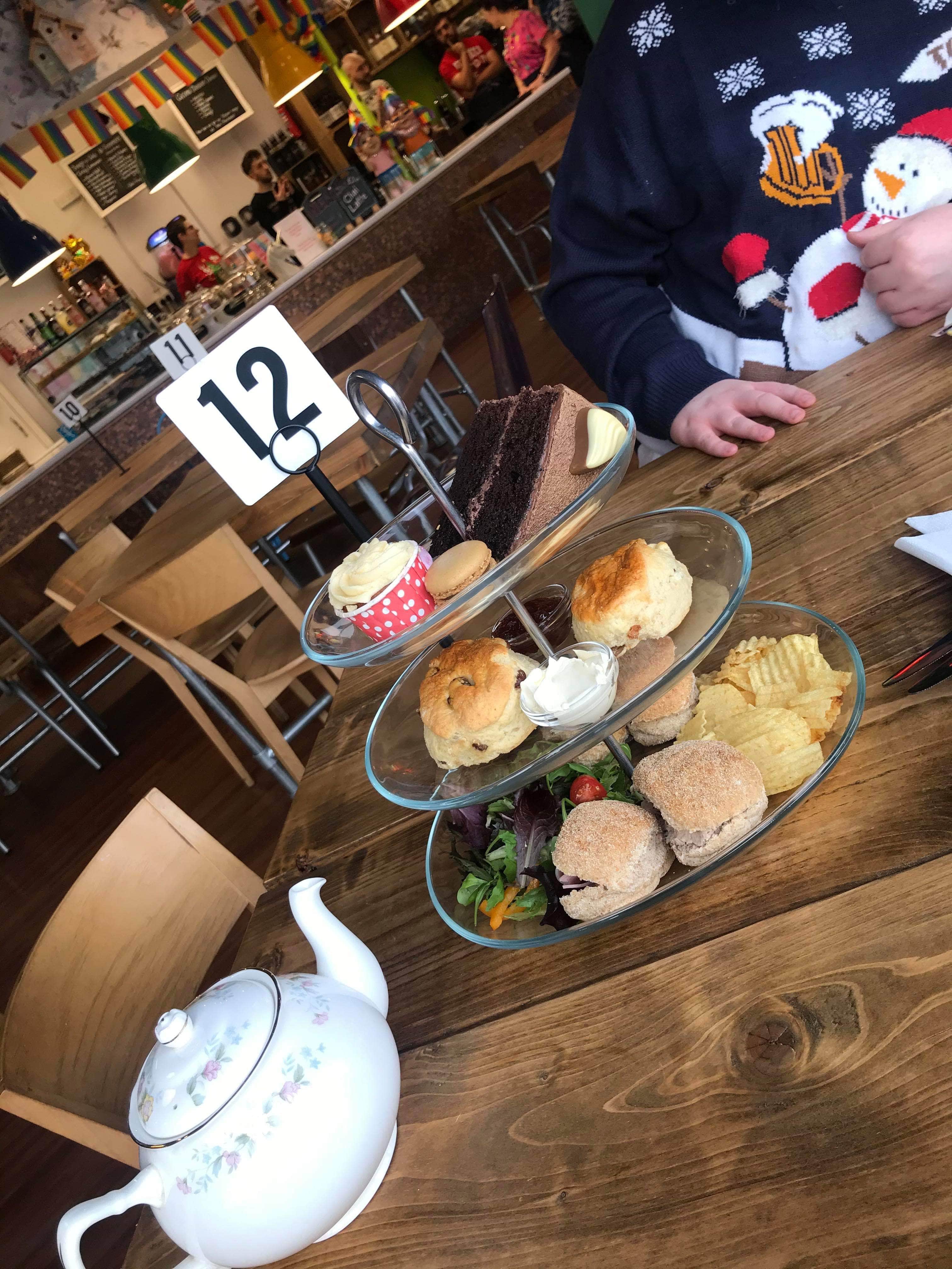 A tall silver metal cake stand that has some white teacake sandwiches, some crisps, a pile of green salad, two large circular golden brown scones, a cylindrical jar full of white clotted cream, a cylindrical jar full of some dark red jam, a large circular chocolate covered cookie, a small circular white macaron, a large circular white cupcake and a large slice of dark brown chocolate cake on it on a large rectangular light wooden table, on a white background.