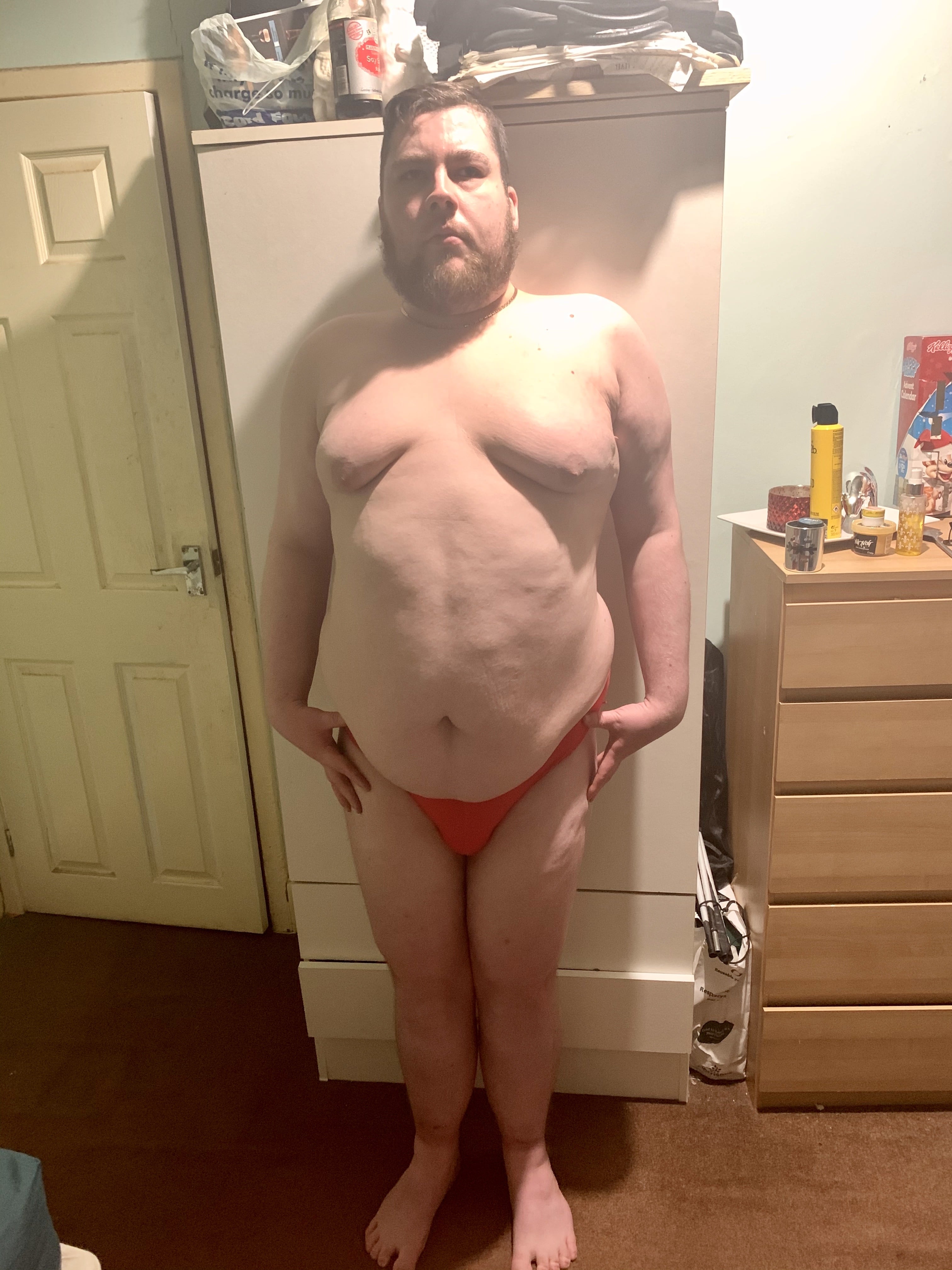 A tall bearded man wearing a pair of bright red briefs with a black waistband that has Box Menswear written in bold white writing on it stood against a light wooden wardrobe.