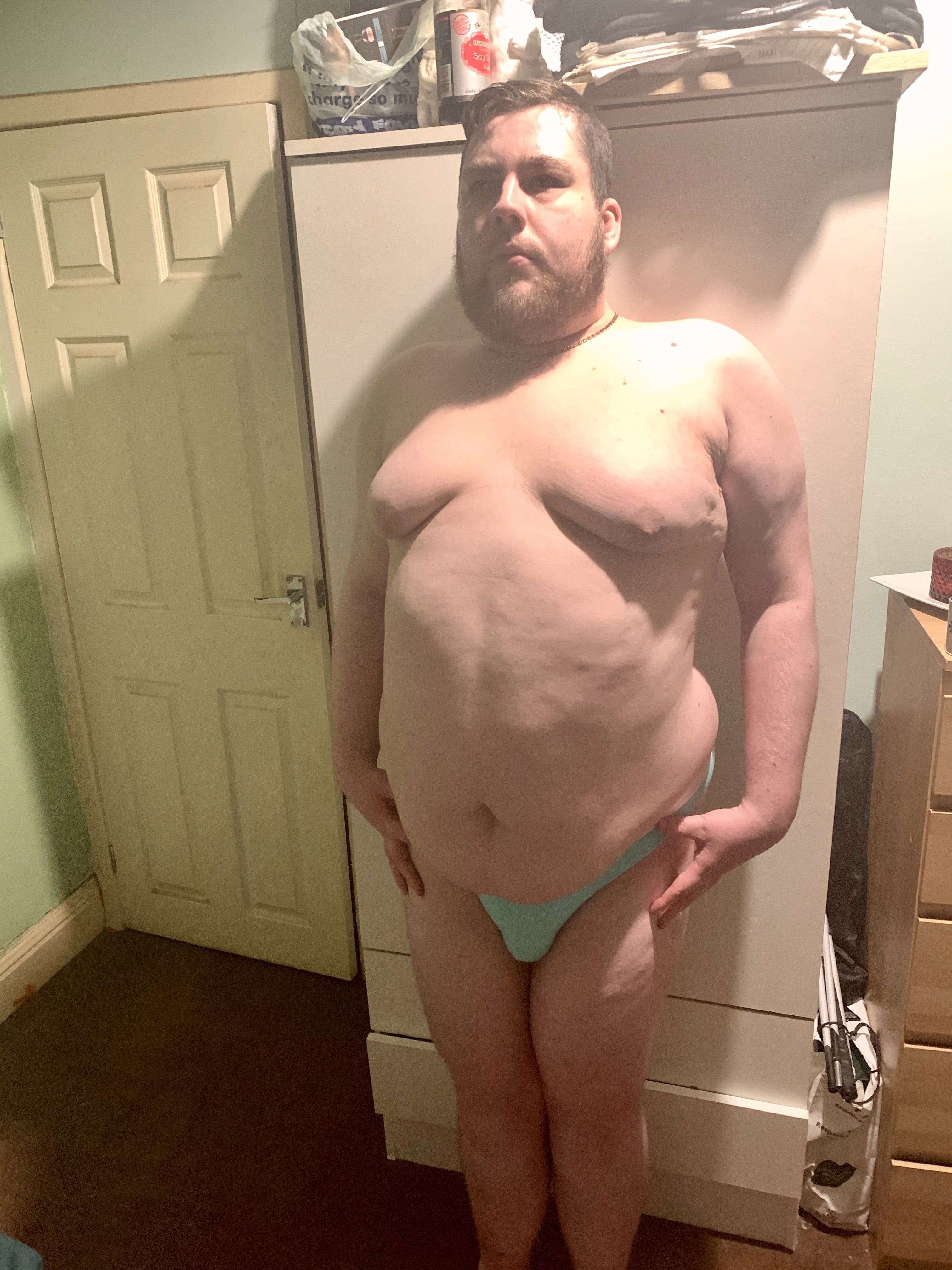 A tall bearded man wearing a pair of bright teal briefs with a black waistband that has Box Menswear written in bold white writing on it stood against a light wooden wardrobe.