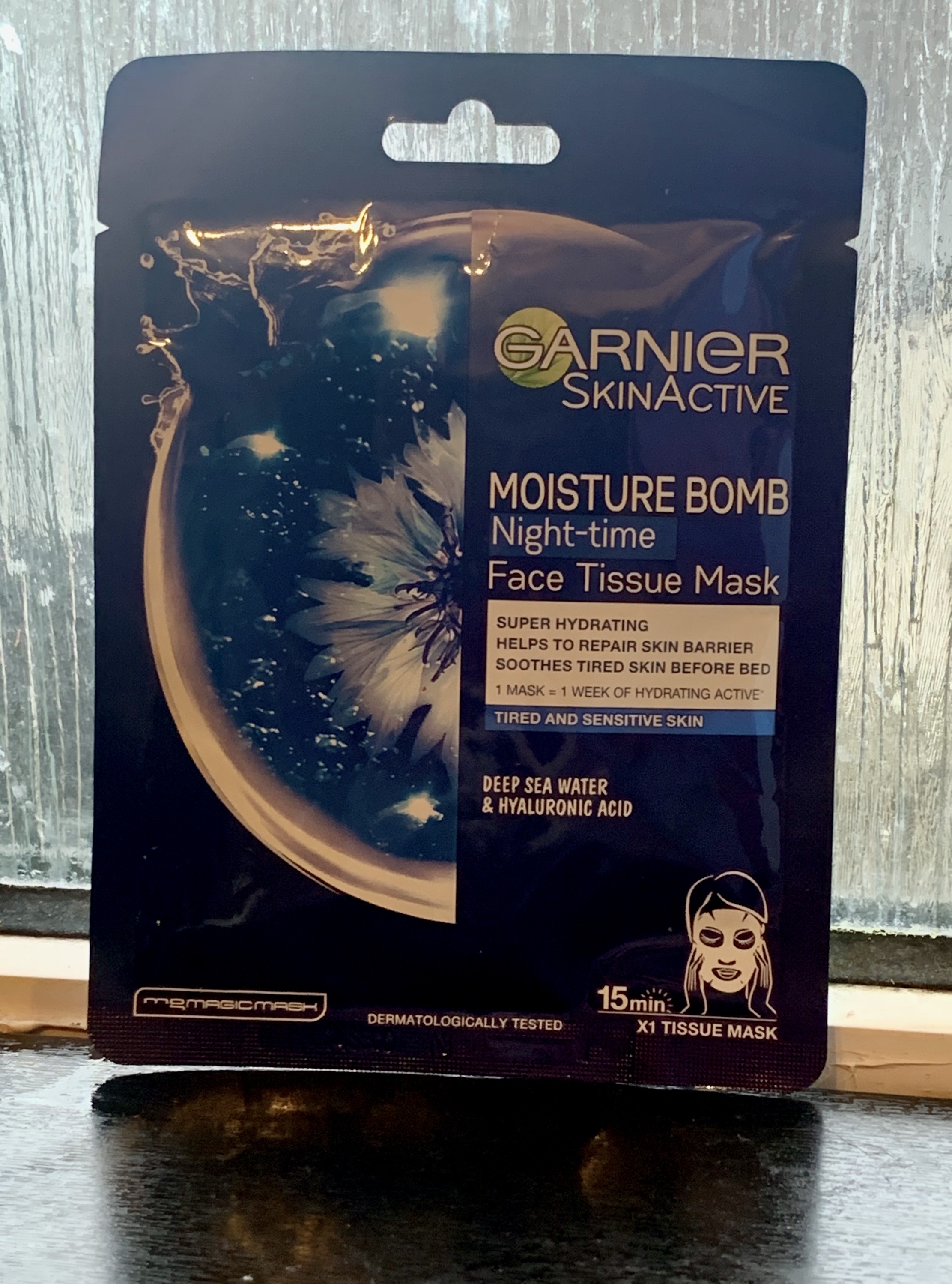Feeling Moist All Night Long | Garnier Moisture Bomb Night Time Face Tissue Mask A tall rectangular black plastic pouch that has Garnier Moisture Bomb Night Time Face Tissue Mask written in bold gold writing with a picture of a droplet of water on it, on a white background.