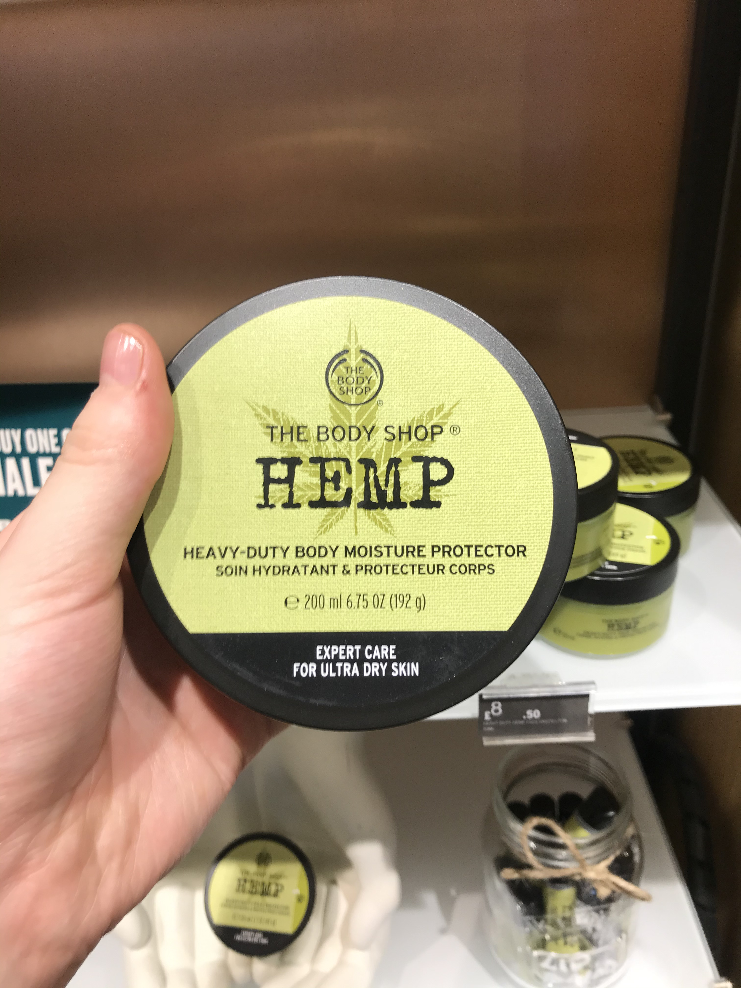 A short and wide cylindrical Black plastic tub with a dark green lid and a bright green label that has The Body Shop Hemp Heavy-duty Body Moisture Protector written in dark green writing on it, on a white background.