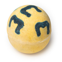 A large spherical shaped bright yellow Bath Bomb that has a dark grey and blue M pattern all over it, on a white background.