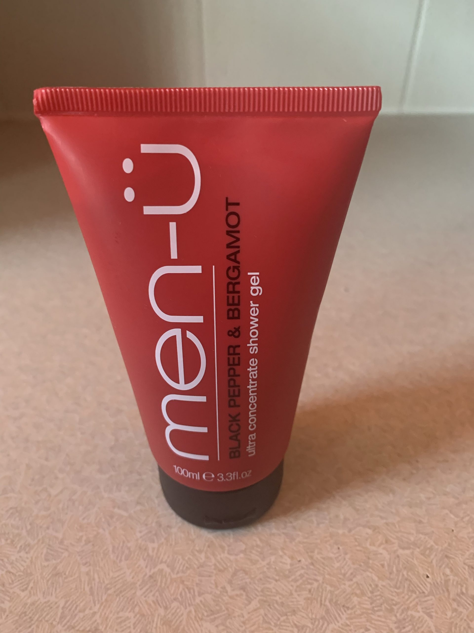 A short conical bright red plastic tube with a black lid and a black label that has Men-ü Shower Gel written in bold white writing and Black Pepper & Bergamot written in bold black writing on it, on a white background.