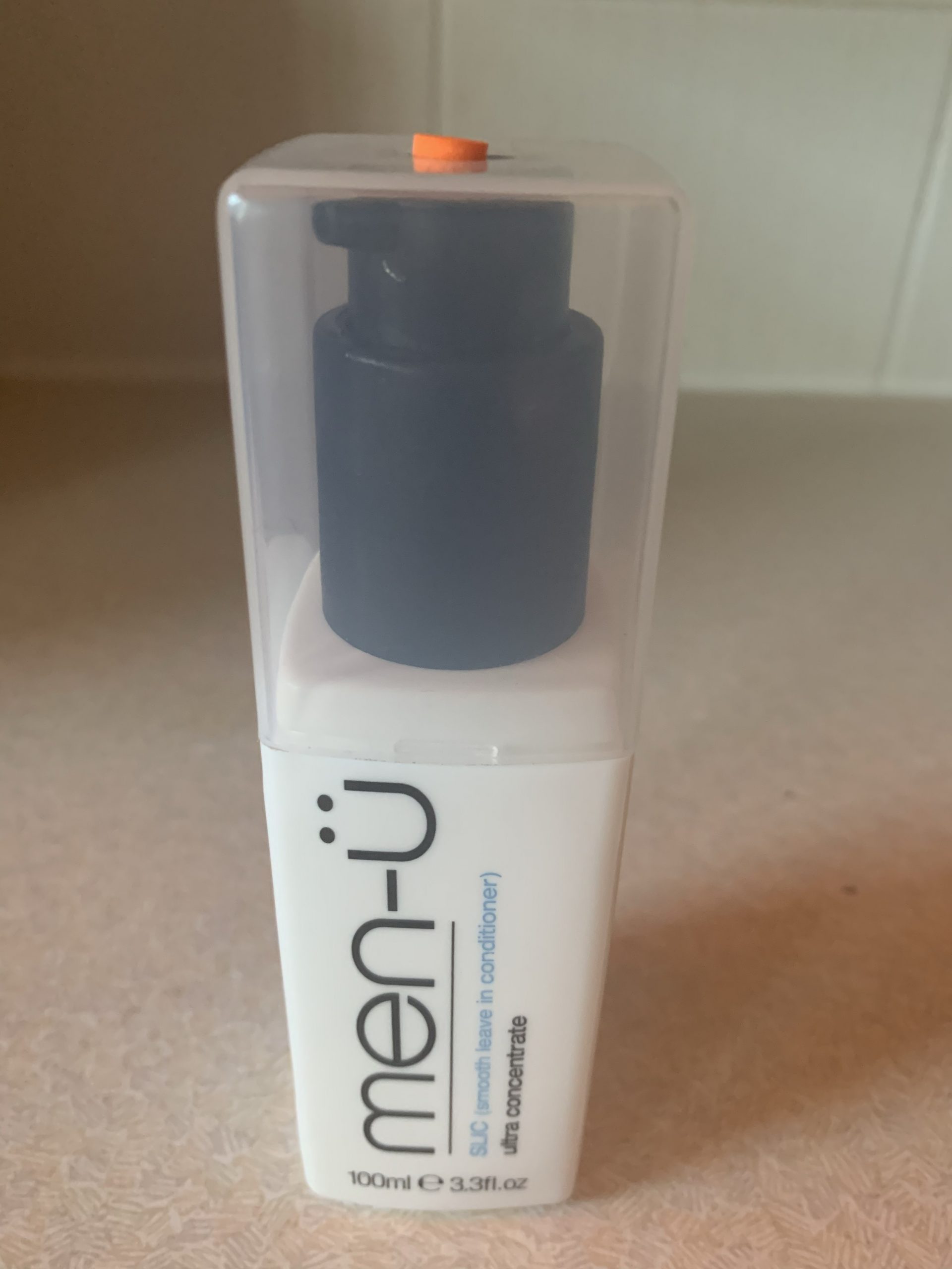 A short rectangular white plastic Bottle with a clear lid that has Men-ü written in bold black writing and Slick, smooth leave in conditioner written in bold blue writing on it, on a white background.