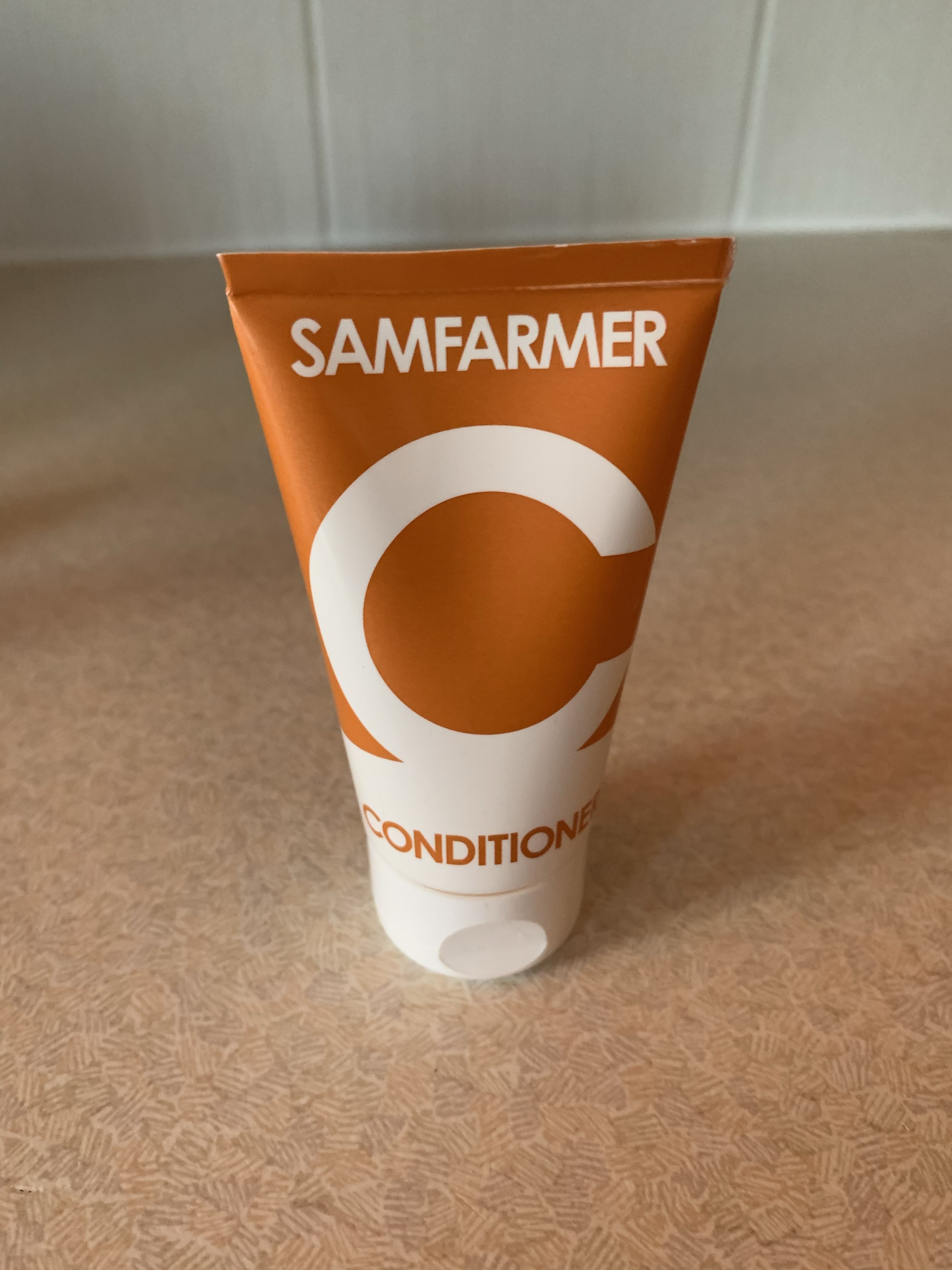 A short flat conical dark purple and white plastic tube that has Sam Farmer written in bold white writing and Face Wash written in dark purple writing on it, on a white background.