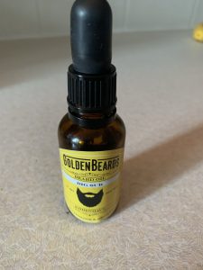 A short cylindrical Brown Glass bottle with a black rubber pipette lid and a bright yellow label that has Golden Beards Big Sur Organic Beard Oil written in bold black writing and a picture of a Beard on it, on a white background.