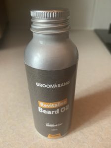 A tall cylindrical silver metallic plastic Bottle with a dark grey label that has Groomarang Beard Oil written in bold white writing on it, on a white background.