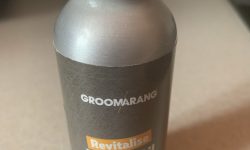 A tall cylindrical silver metallic plastic Bottle with a dark grey label that has Groomarang Beard Oil written in bold white writing on it, on a white background.