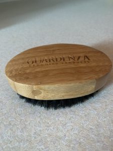 A large oval light wooden beard brush with black bristles that has Guardenza engraved on top of it, on a white background.