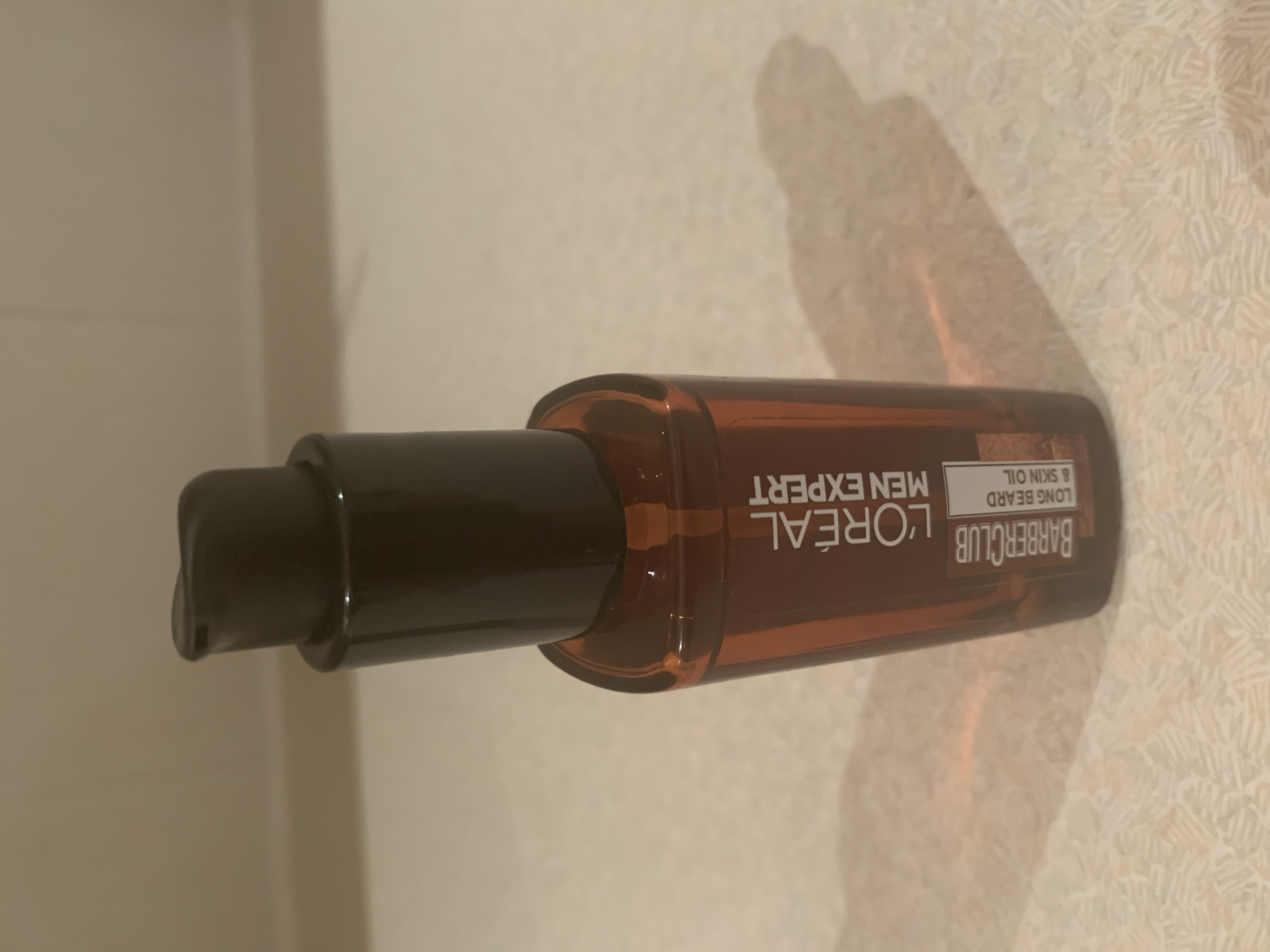 A short rectangular glass bottle full of some dark brown liquid with a black plastic pump lid and a black label that has L'Oreal Paris Men Expert Barber Club Long Beard & Skin Oil written in bold white writing on it, on a white background.