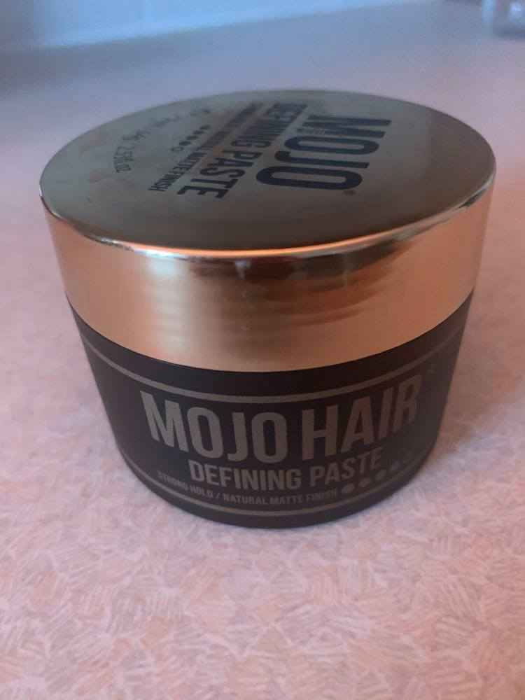 Putting The Mojo Back Into My Hair | Mojo Hair* Styling Products | Gifted A short cylindrical matte black plastic tub with a glossy gold plastic lid that has Mojo Hair* Defining Paste written in bold gold writing on it, on a white background.