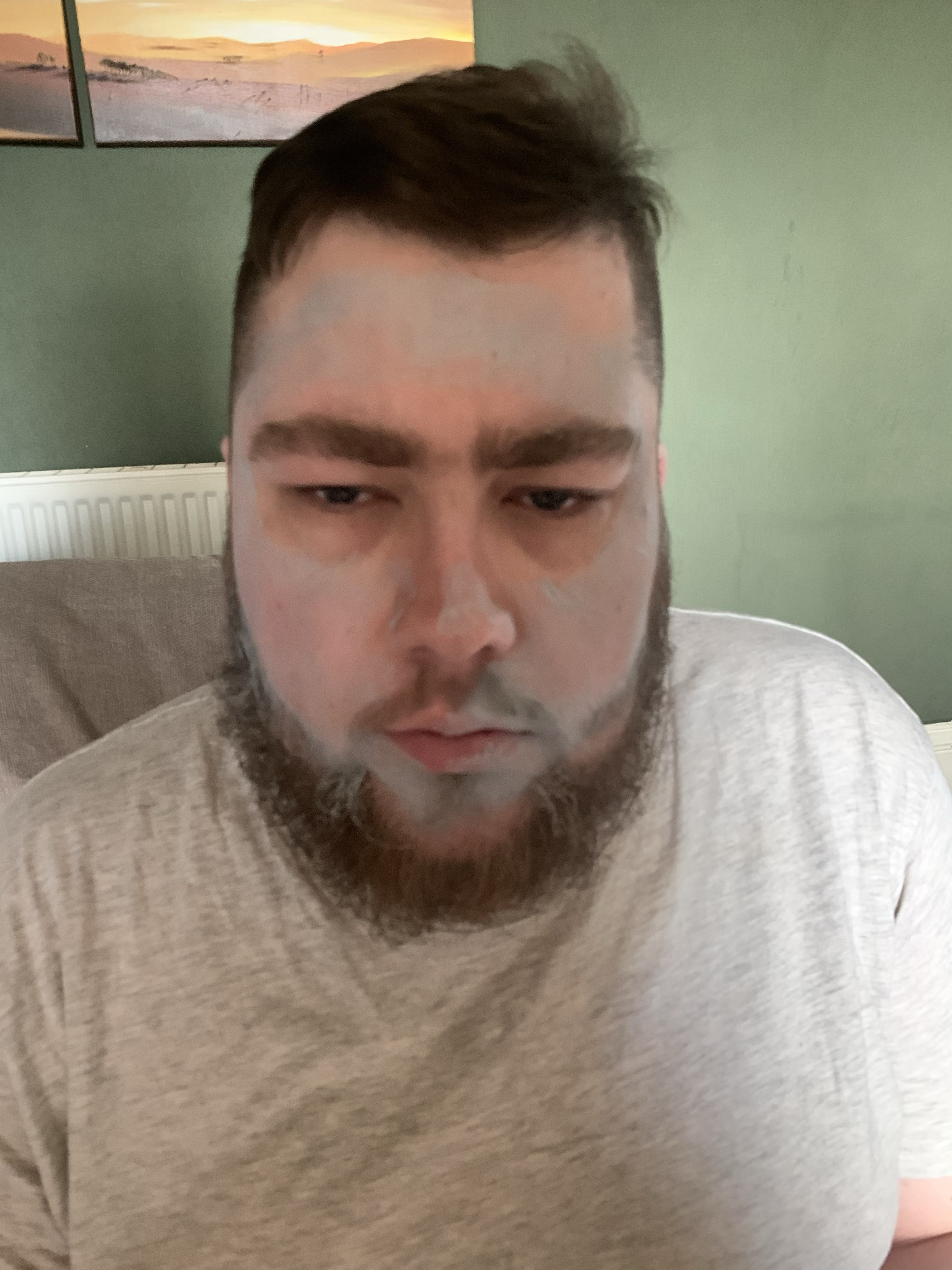A bearded man that has some creamy pale green face mask all over his face sat on a dark blue sofa.