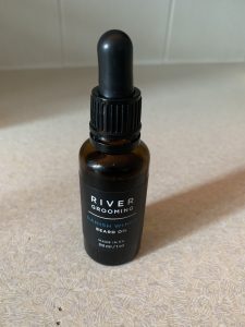 A short and thin cylindrical Brown Glass Bottle with a Black Rubber Pipette Lid and a black label that has River Grooming Danish Winds Beard Oil written in bold white writing on it, on a white background.