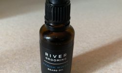 A short and thin cylindrical Brown Glass Bottle with a Black Rubber Pipette Lid and a black label that has River Grooming Danish Winds Beard Oil written in bold white writing on it, on a white background.