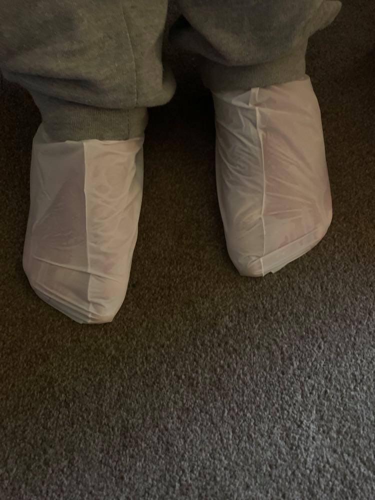 Take The Dry Skin Off My Feet | Superdrug Tea Tree & Peppermint Nourishing Foot Mask Me wearing a pair of bright white plastic socks stood on a dark grey carpet.