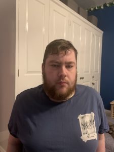 A tall Bearded Man wearing a dark blue t-shirt stood against a white wardrobe.