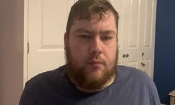 A tall Bearded Man wearing a dark blue t-shirt stood against a white wardrobe.